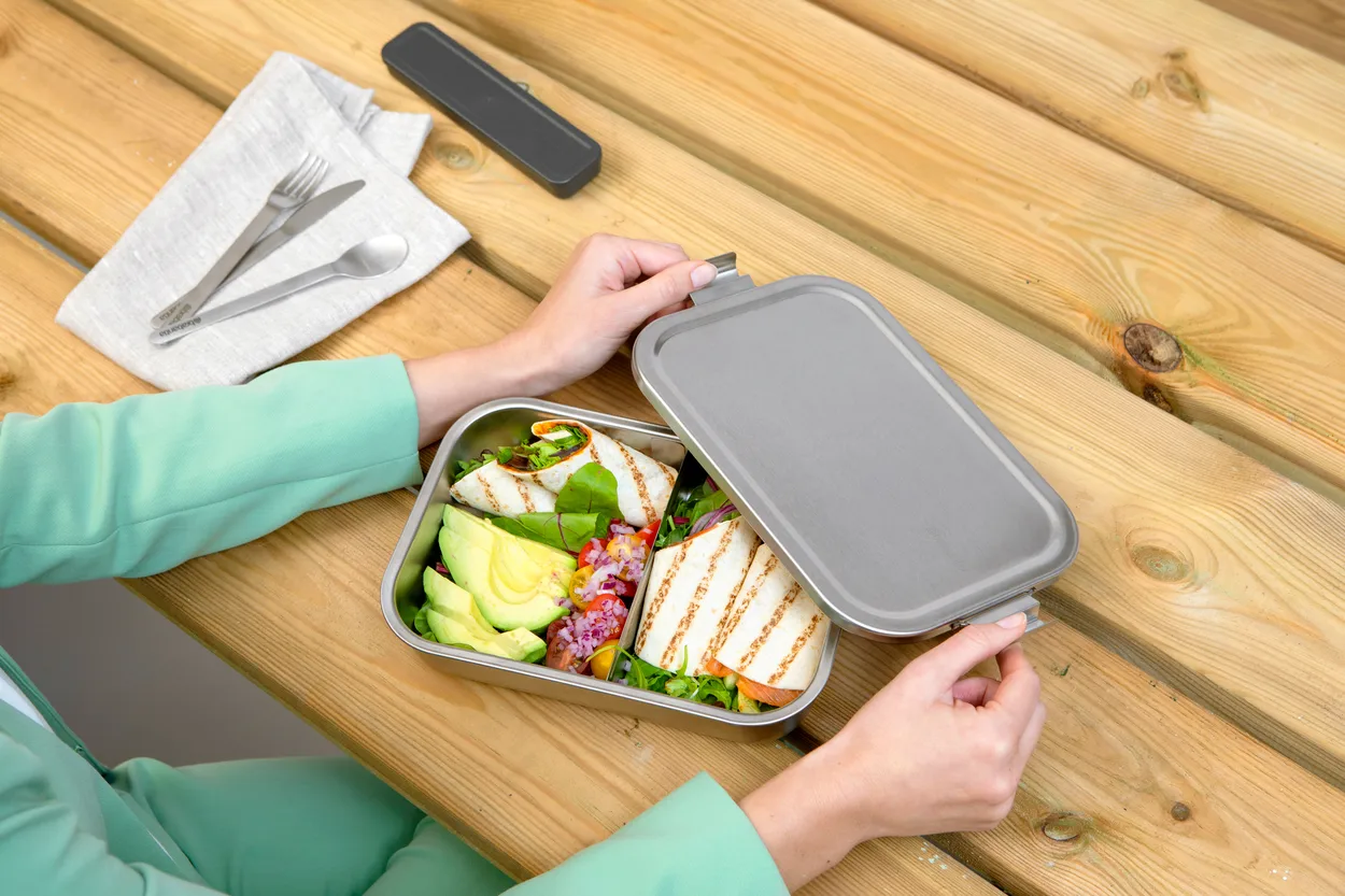 To hænder løfter det grå låg af en åben madkasse i stål på et træbord, hvor der ligger grillet ost, avocado, salat og grøntsager i to rum, og ved siden af en foldet serviet med bestik