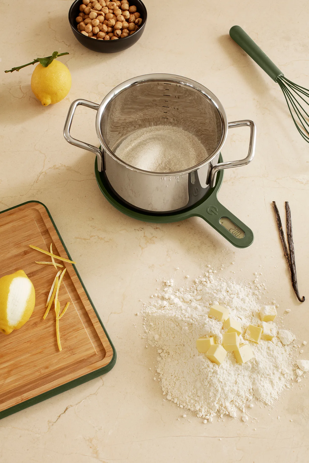 Rustfri kasserolle med mel står oven på en mørkegrøn køkkenvægt på en cremefarvet bordplade, med skærebræt, citronskal, smørterninger og vaniljestænger ved siden af