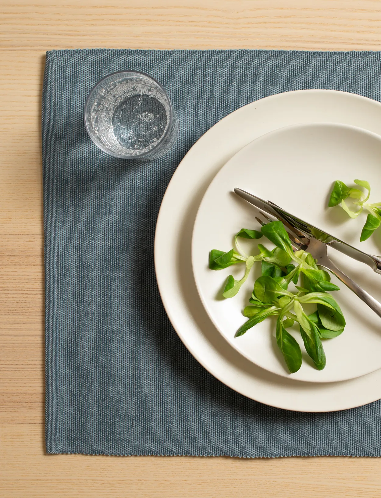 Blågrå dækkeserviet på et lyst træbord med en creme tallerken, bestik og friske salatblade