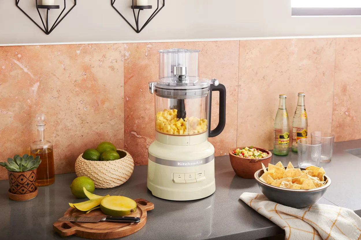 Cremefarvet foodprocessor med majs i skålen på en køkkenbordplade med lime, citron og guacamole