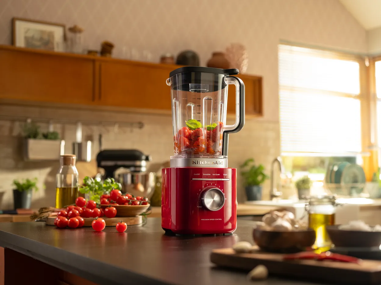 Rød blender på en mørk bordplade i et køkken med jordbær og mynte i den klare kande, en skål jordbær, en kurv cocktailtomater og en oliekande ved siden af