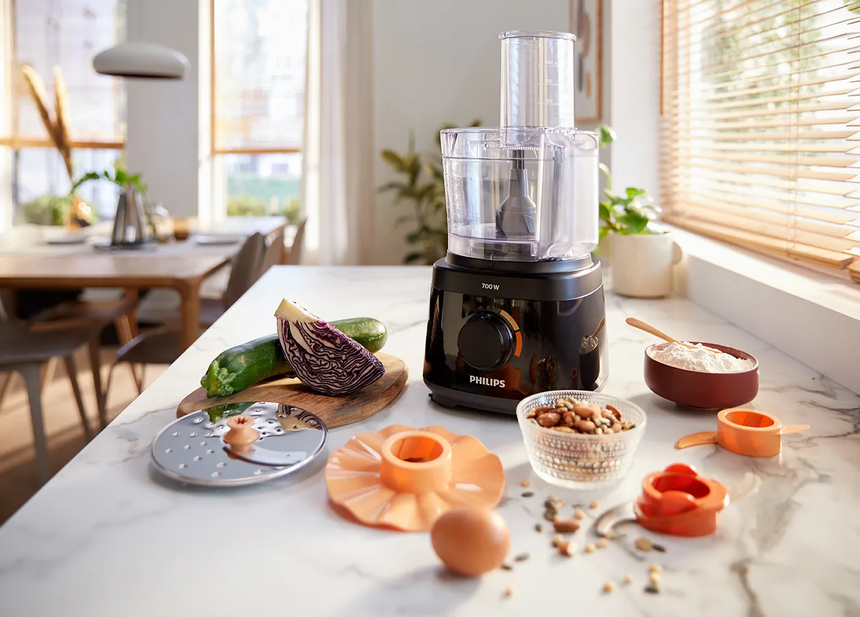 Sort foodprocessor på en hvid marmorbordplade ved vinduet med en rivejernsskive, et bræt med squash og rødkål, en skål nødder og en skål mel foran