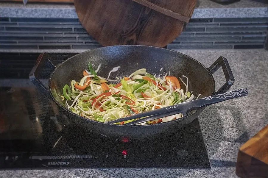 Wok med snittede grøntsager på en sort induktionsplade ved siden af et skærebræt