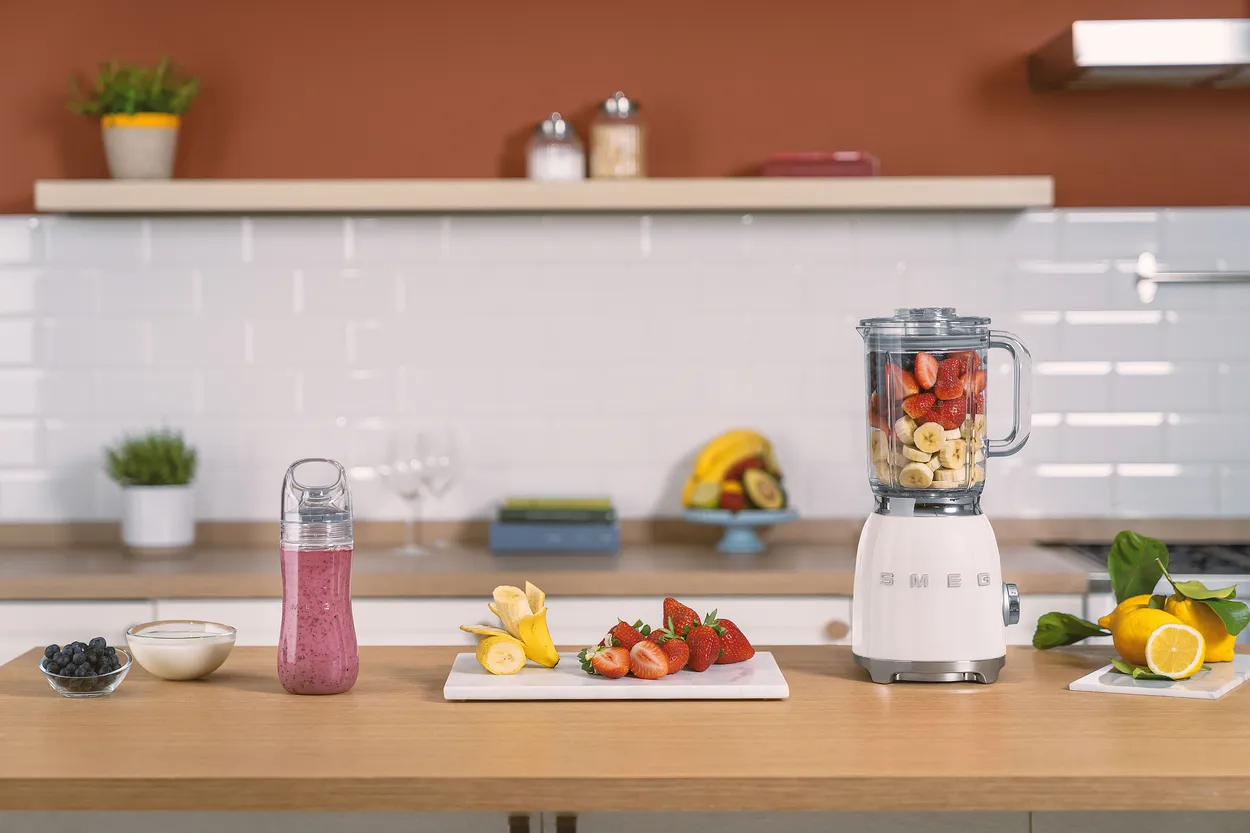 Hvid blender på en træbordplade med halve jordbær og bananskiver i kanden, en flaske med rød smoothie og et skærebræt med frugt ved siden af