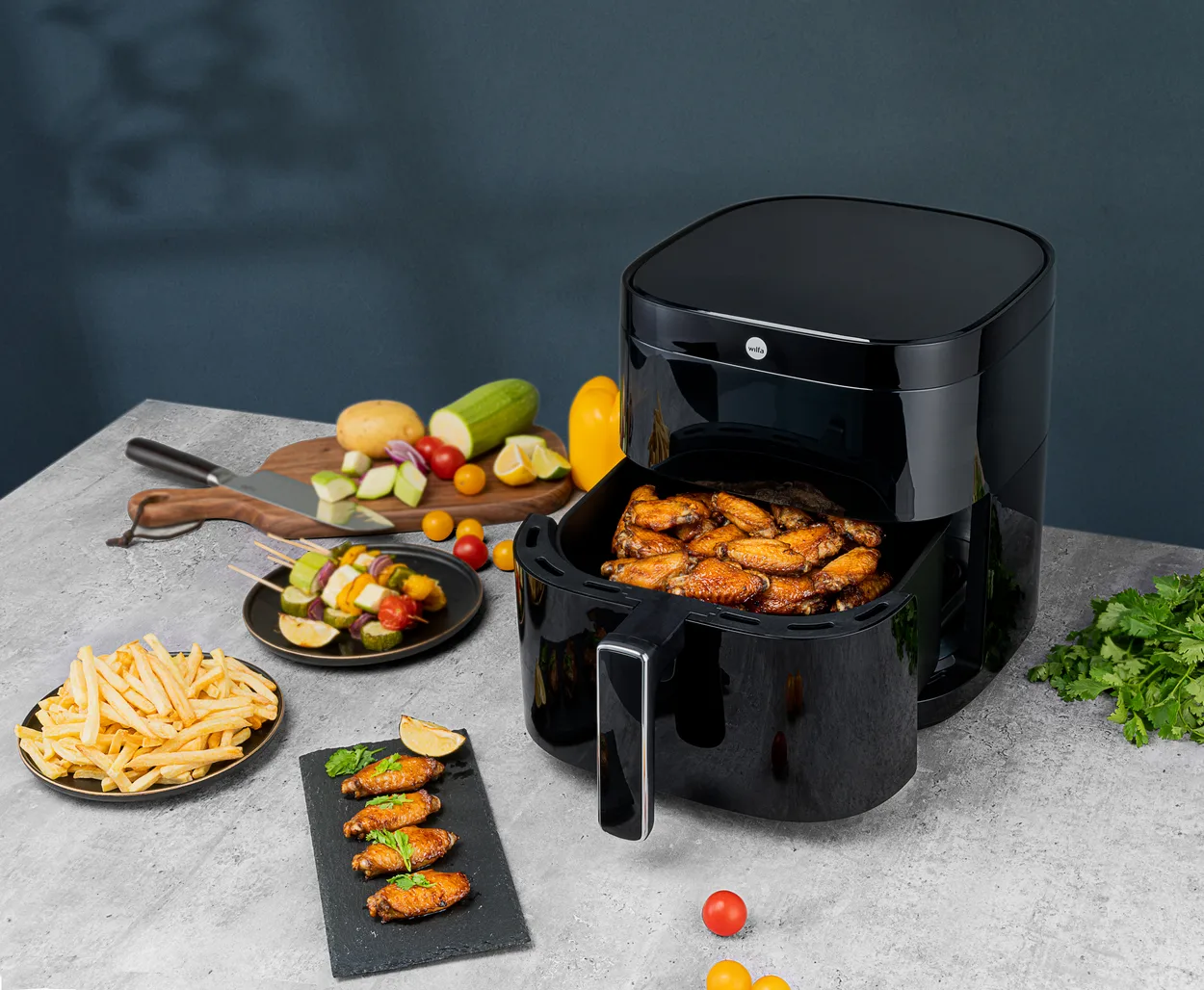 Sort airfryer på en grå stenbordplade foran en mørkeblå væg med skuffen trukket ud fuld af glaserede kyllingevinger, og omkring den et fad pommes frites, to grøntsagsspyd og et træbræt med kartoffel, majs og tomater