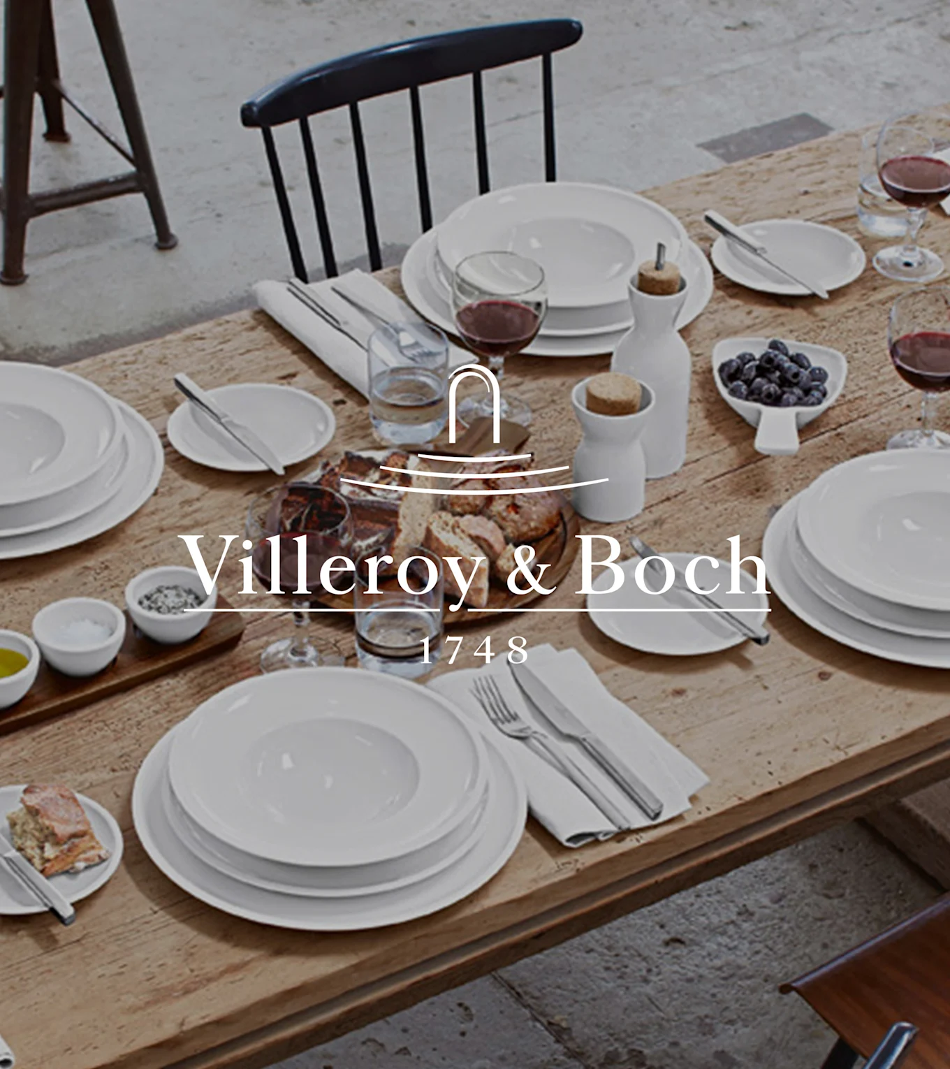Et rustikt træbord dækket med Villeroy & Boch hvidt stel, vin, brød, blåbær, salt, peber og olivenolie til et måltid.