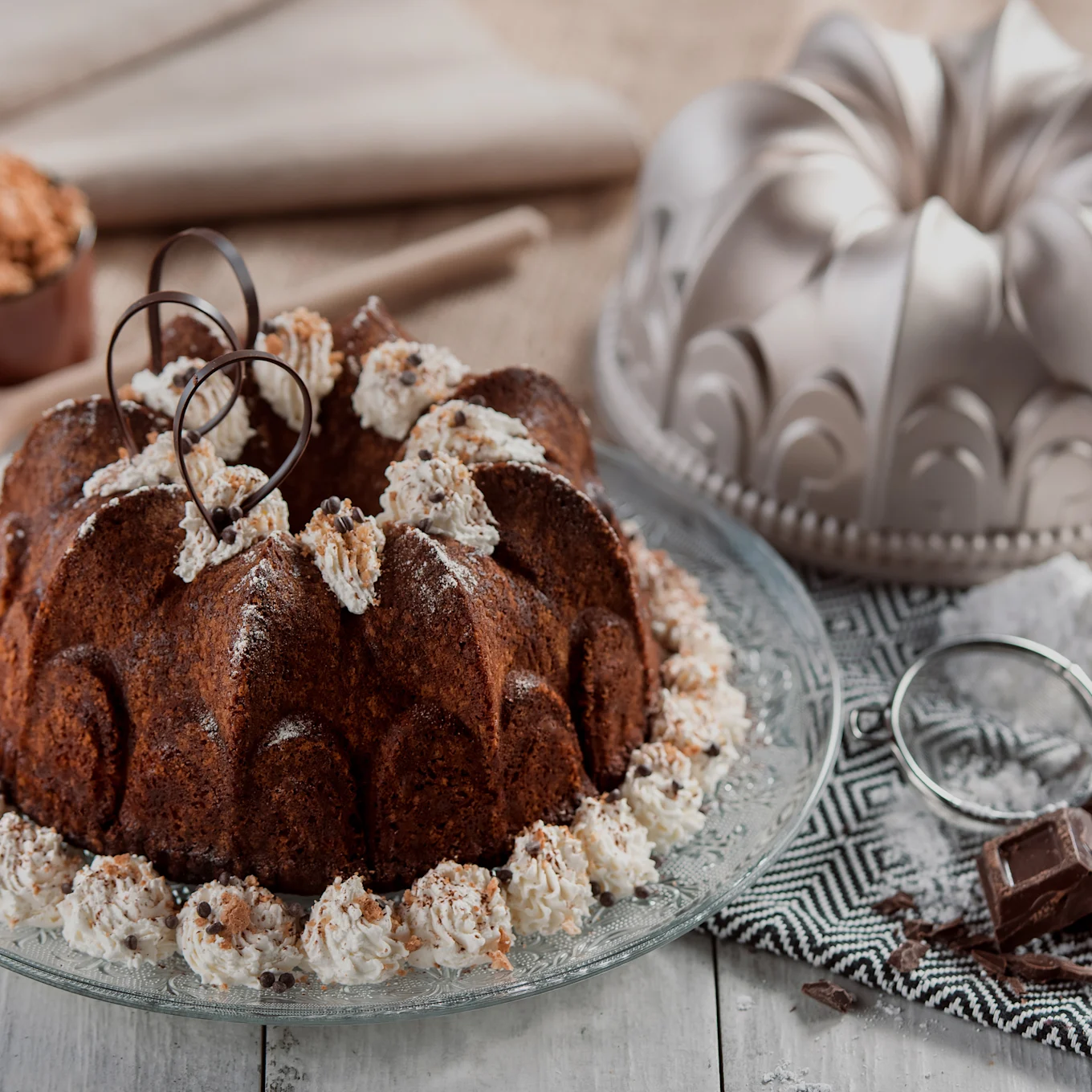 Chokolade-bundtkage med flødeskum, chokoladestykker og chokoladekrøller på en glasplade, med en bundtform og ingredienser i baggrunden.