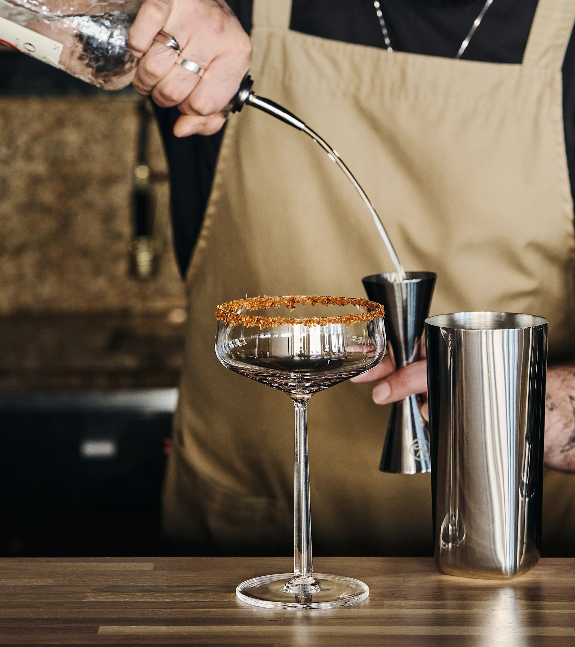 En bartender hælder en drink fra en flaske i et måleglas, med et sukkerkantet cocktailglas og en shaker på en trædisk.