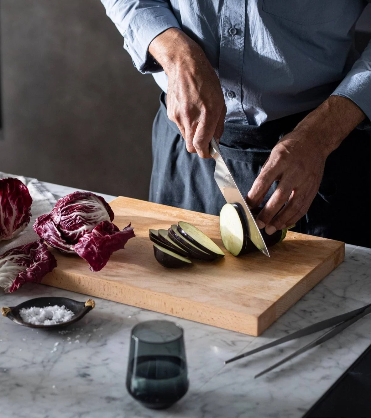 Nærbillede af en person, der skærer aubergine på et træskærebræt med radicchio og salt i nærheden.