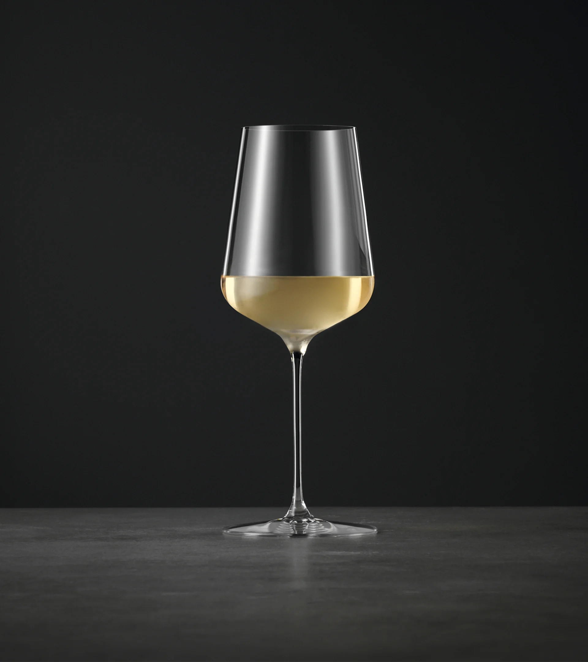 Et højt, elegant vinglas med lang stilk, halvt fyldt med lys hvidvin, mod en mørk baggrund.