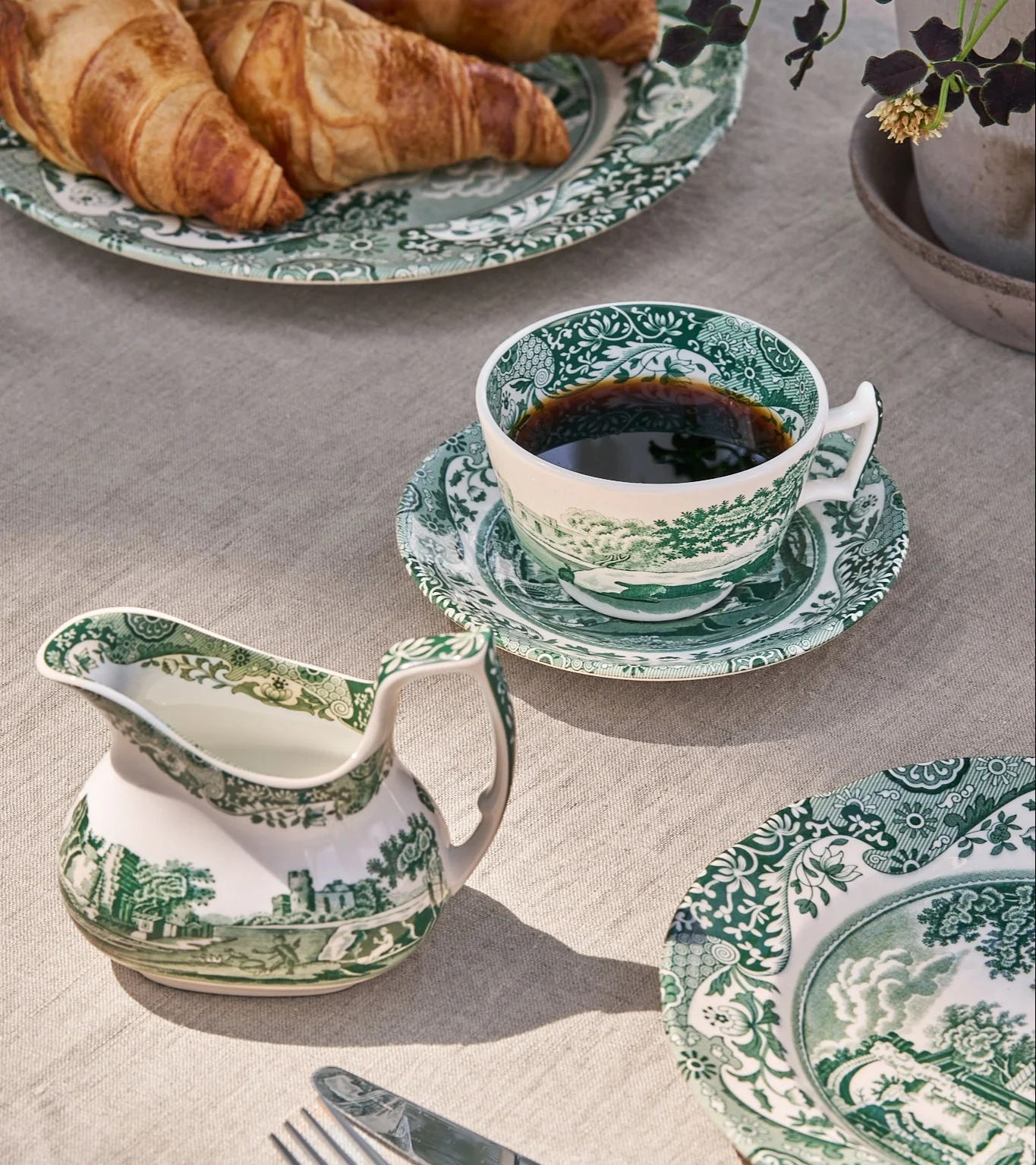 Morgenmadsbord med grøn-hvid mønstret porcelænskop med kaffe, flødekande og tallerkner med croissanter.