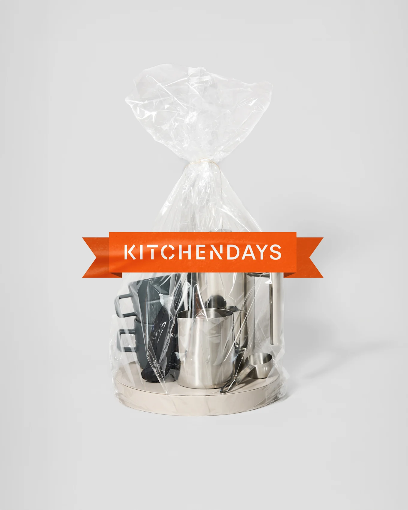 En gennemsigtig gavepose bundet med snor, indeholdende en sølvfarvet kaffemaskine, en sølvfarvet mælkekande og sorte krus på en lys bakke, med et orange "KITCHENDAYS" banner.