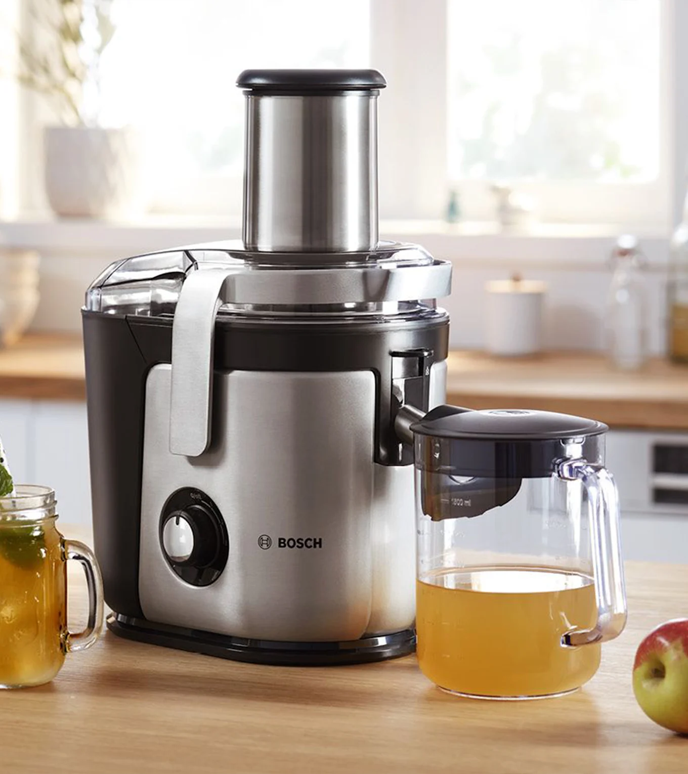 Bosch saftpresser, sølv og sort, på et køkkenbord med en kande æblejuice og et æble.