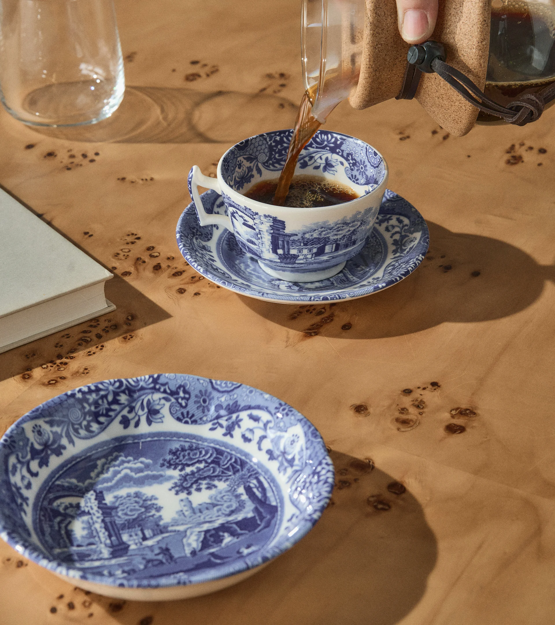 En hånd hælder kaffe i en blå og hvid mønstret tekop på et træbord med en matchende skål, en bog og et glas.