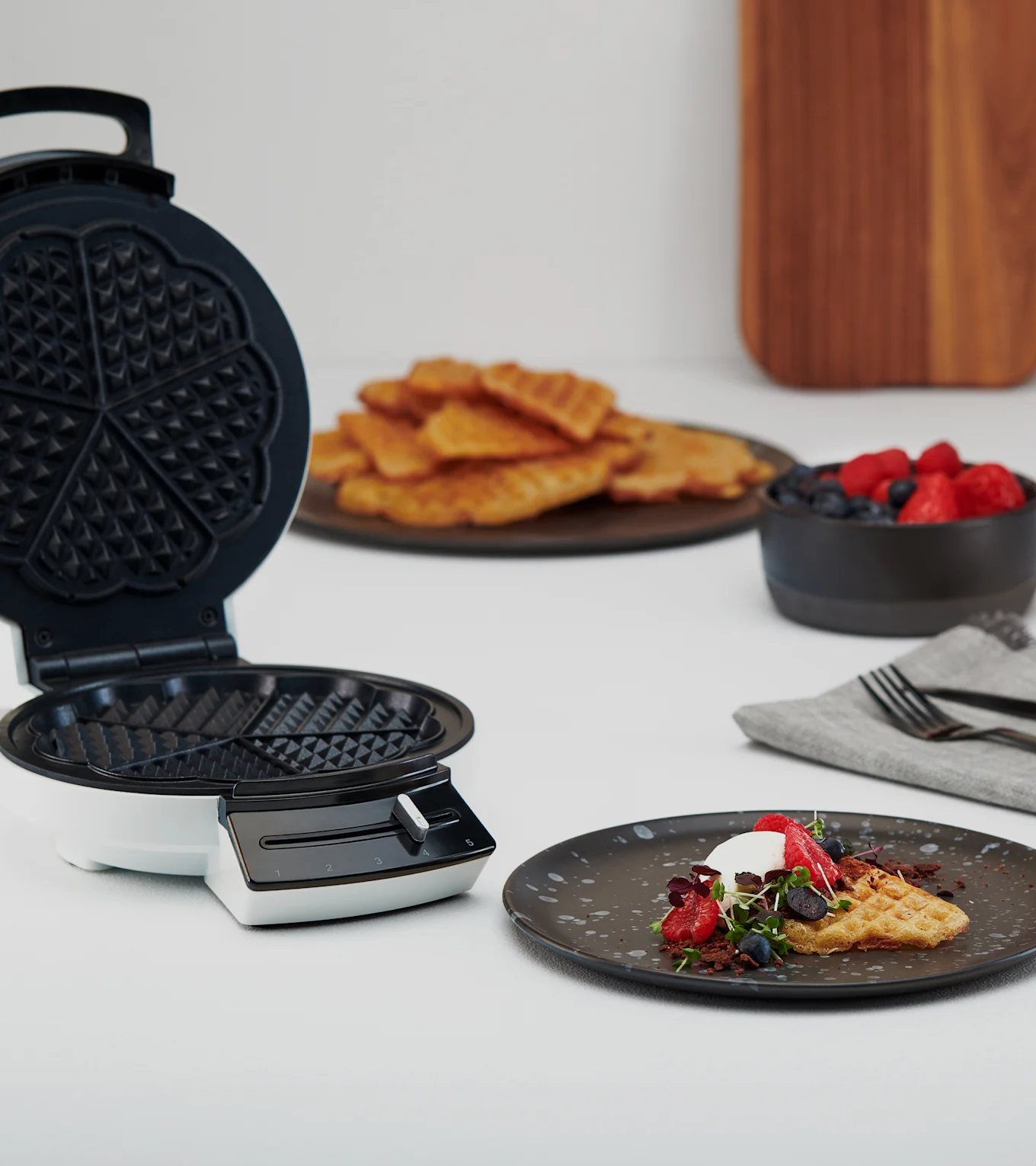 Åbent vaffeljern ved siden af tallerken med vaffel, flødeskum, bær og mikrogrønt. Flere vafler og bær i baggrunden.