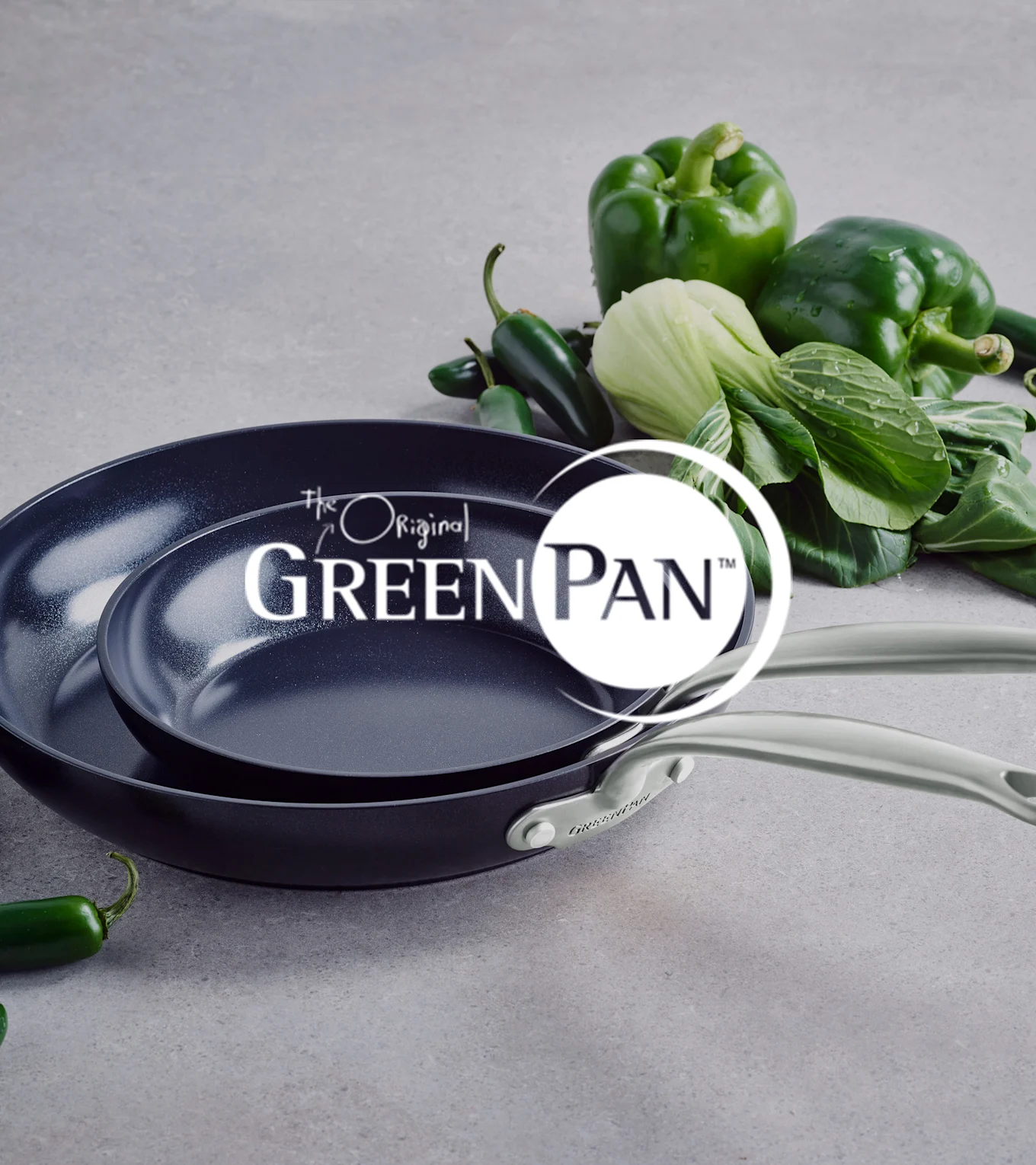 To mørke non-stick pander med lyse håndtag, stablet, ved siden af grøn peberfrugt, pak choi og jalapeños. GreenPan logo.