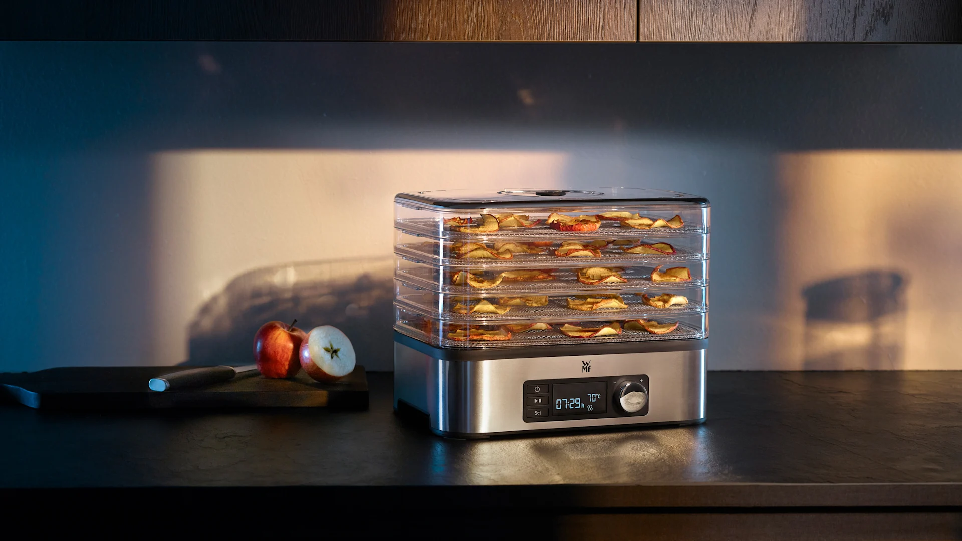 Fødevaredehydrator med klare bakker fyldt med æbleskiver på et mørkt køkkenbord, med friske æbler i nærheden.