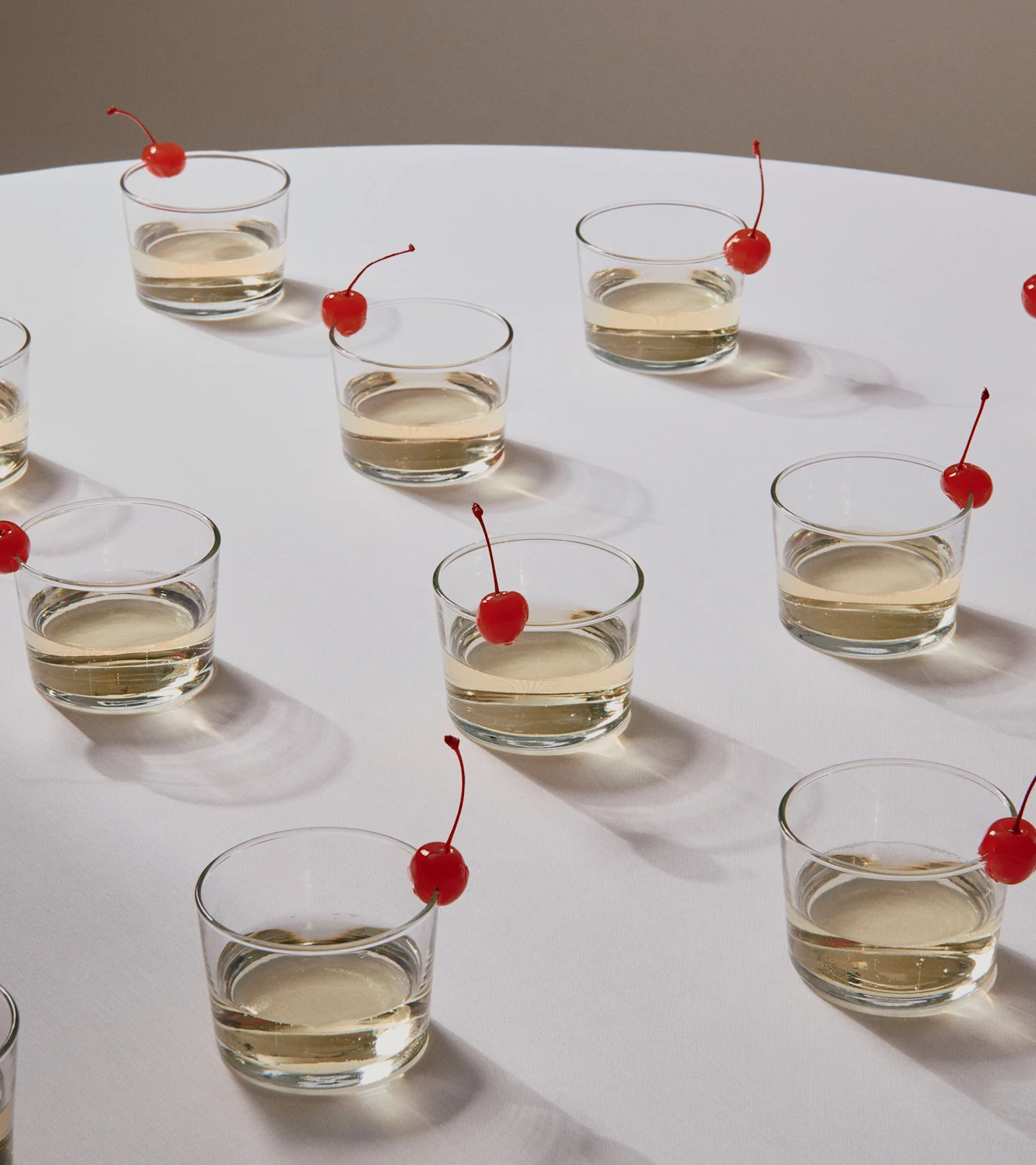 Flere glas med lyse drinks, hver med et rødt kirsebærpynt, på en hvid overflade.