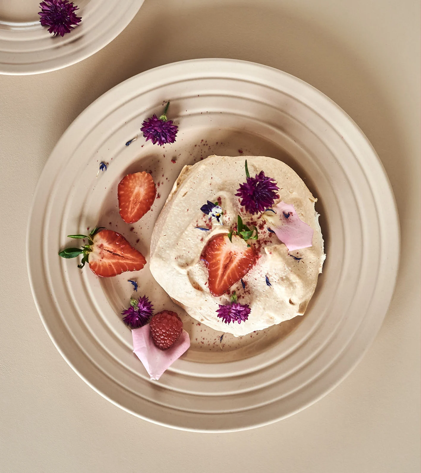 En cremet dessert med jordbær, et hindbær og lilla blomster på en beige riflet tallerken.
