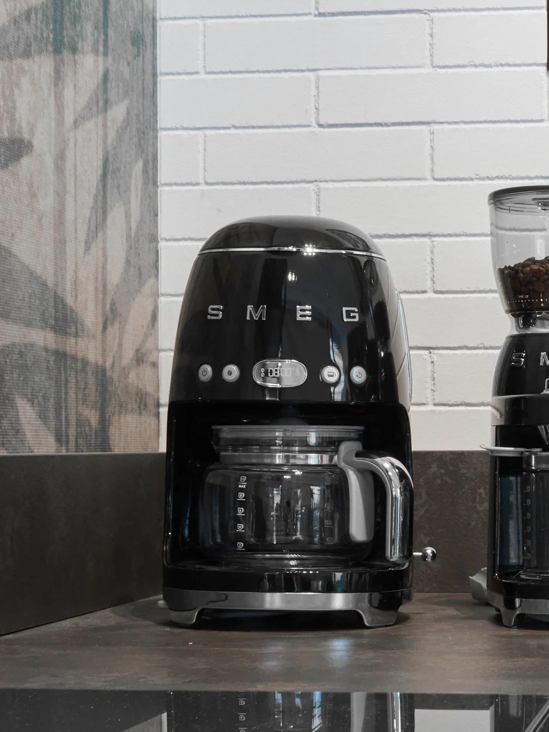 Sort Smeg kaffemaskine med glaskande og digitalt display, der viser '08:00', på en mørk bordplade.