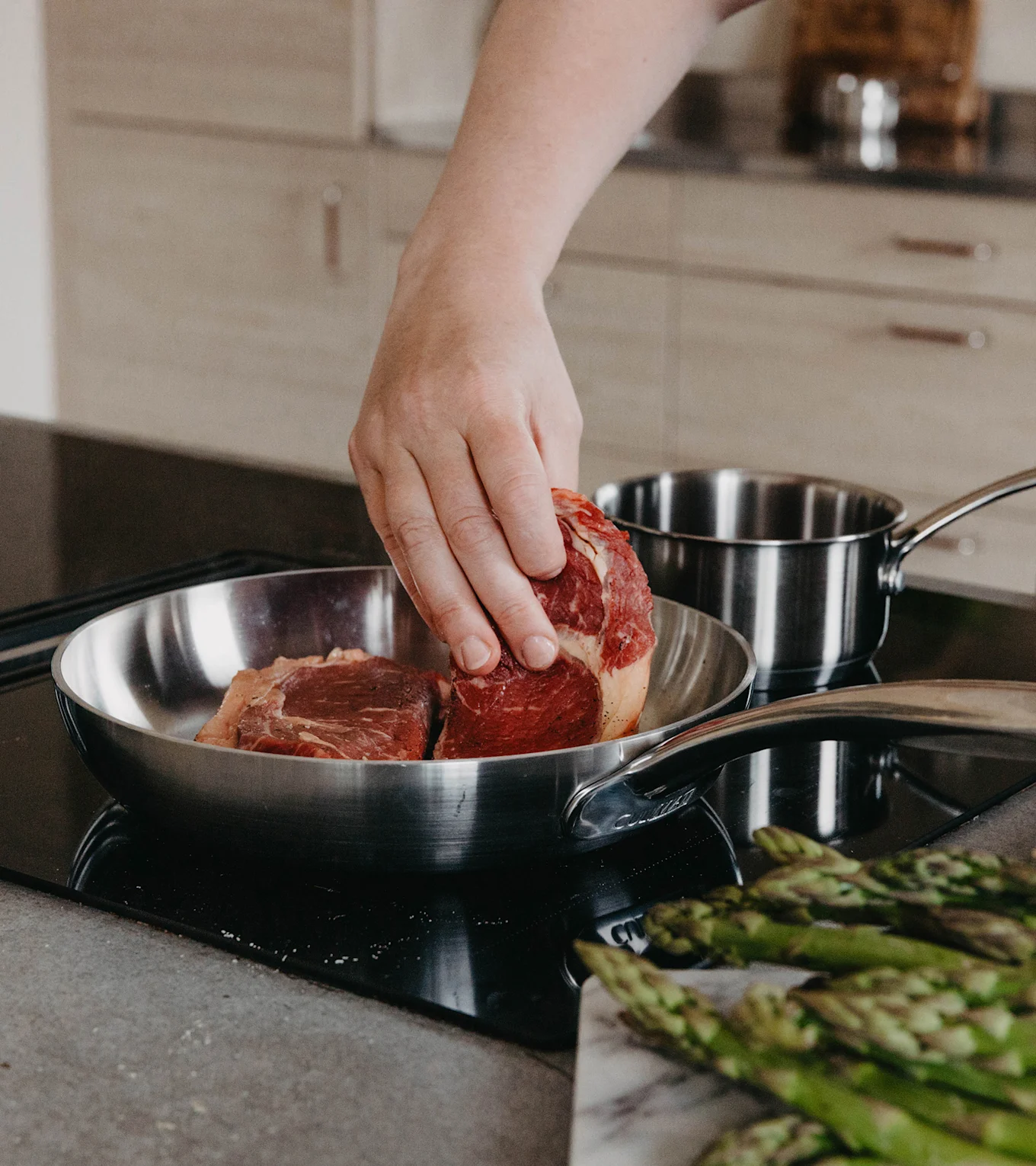 Hånd lægger rå bøf i stegepande af rustfrit stål på kogeplade, med asparges på marmorbræt i forgrunden.