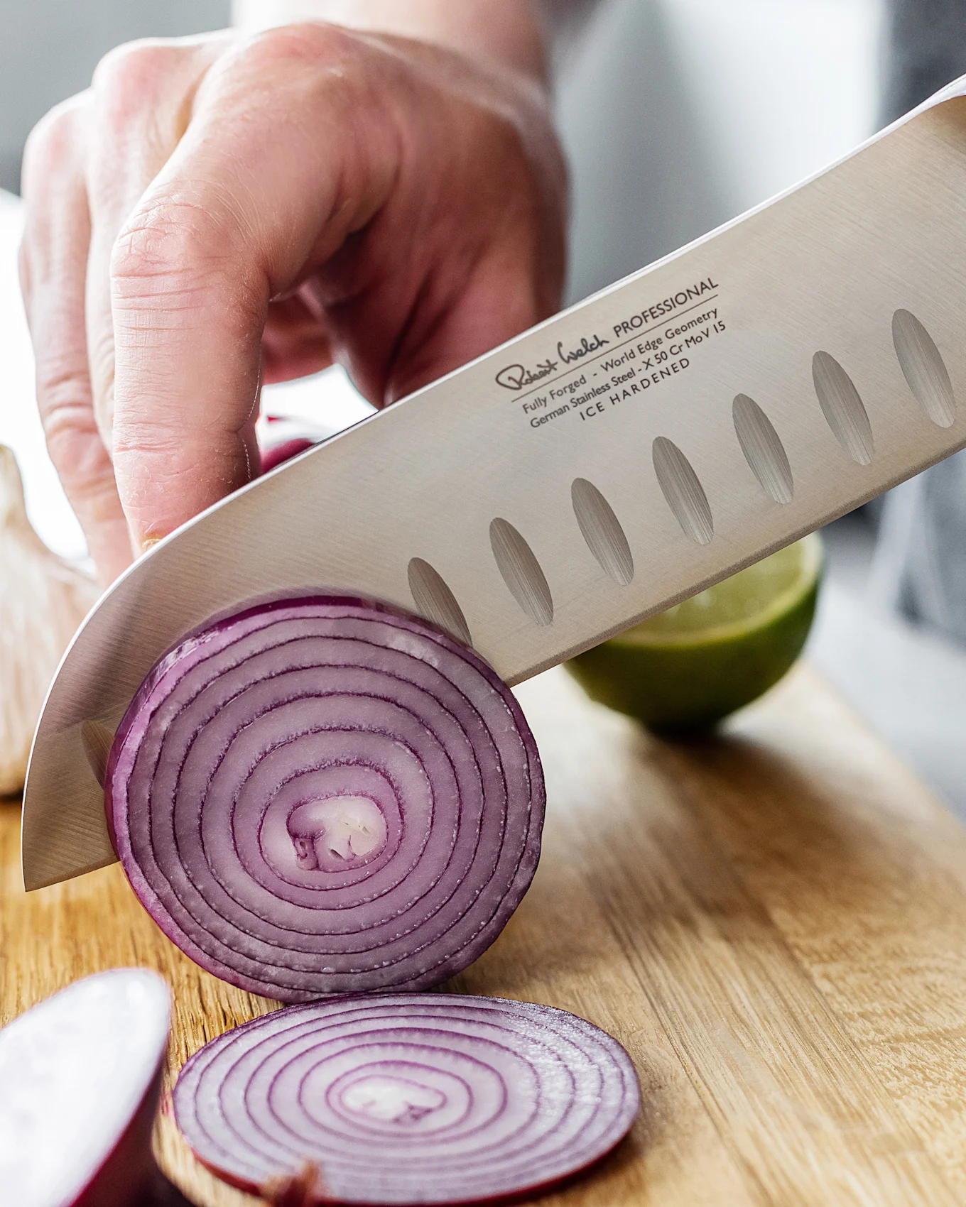 En kokkekniv med tekstureret blad skærer et rødløg på et træbræt, med en lime i baggrunden.