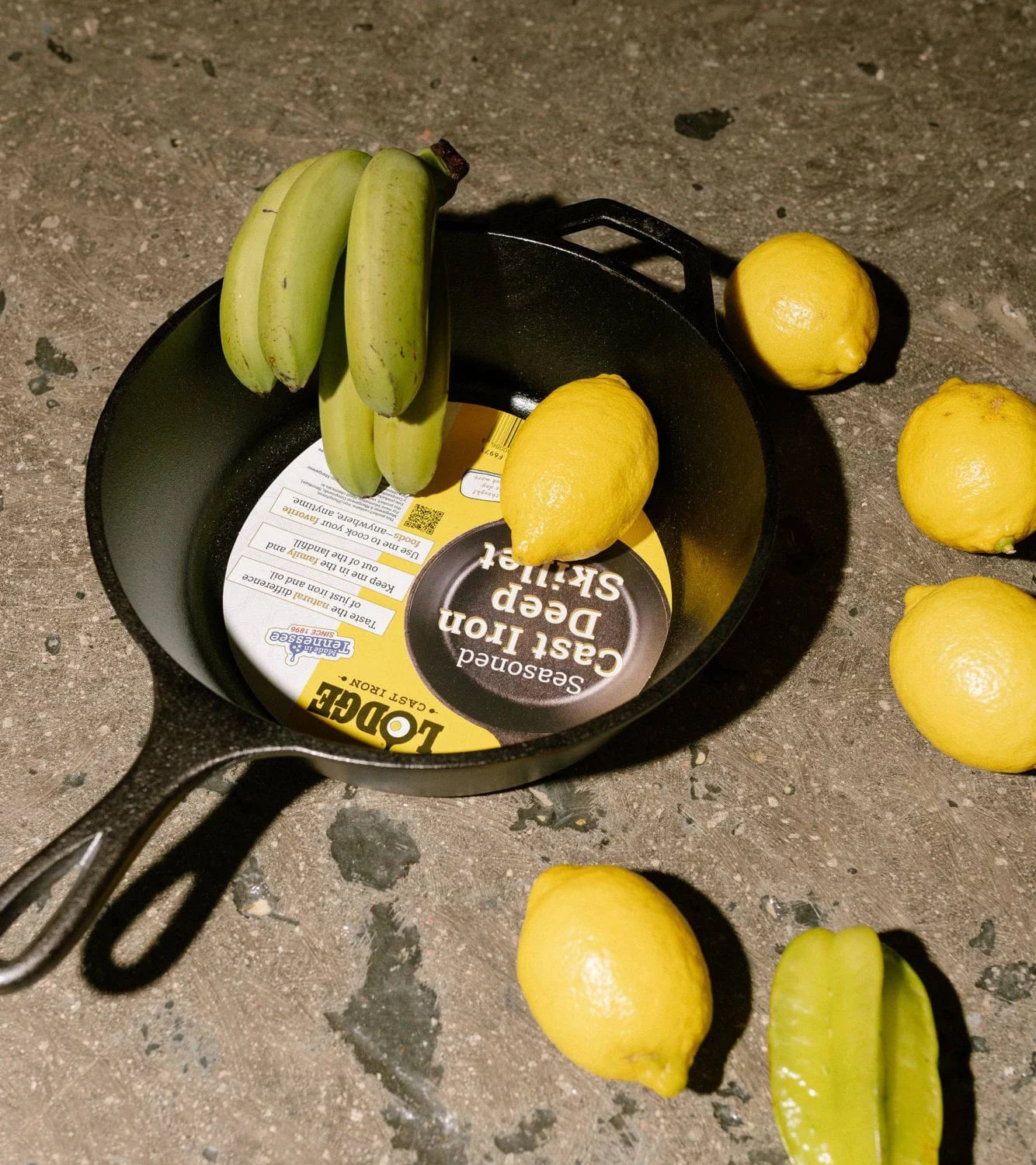 Støbejernspande med et mærkat indeni, umodne bananer, citroner og en stjernefrugt på en mørk overflade.