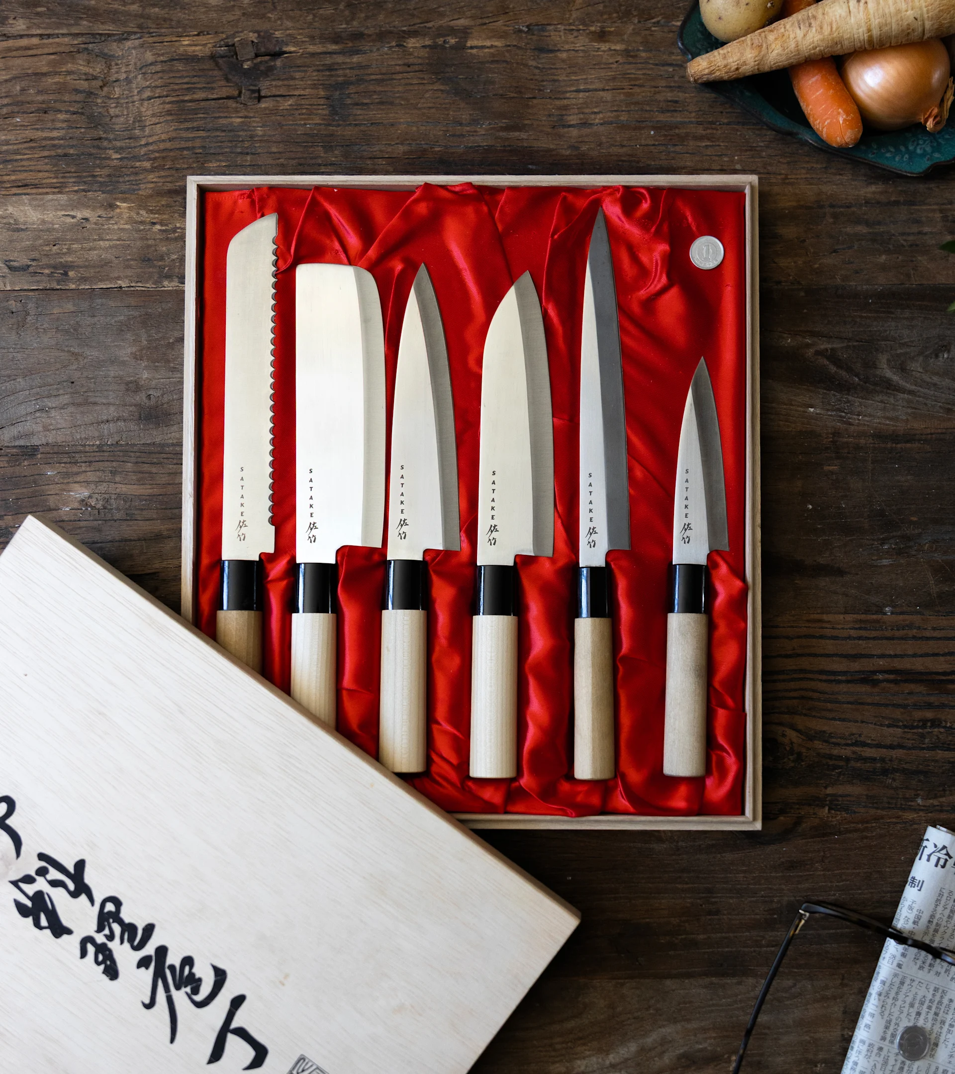 Seks japanske knive med træhåndtag i en rødfôret æske på et træbord, med grøntsager og avis i baggrunden.