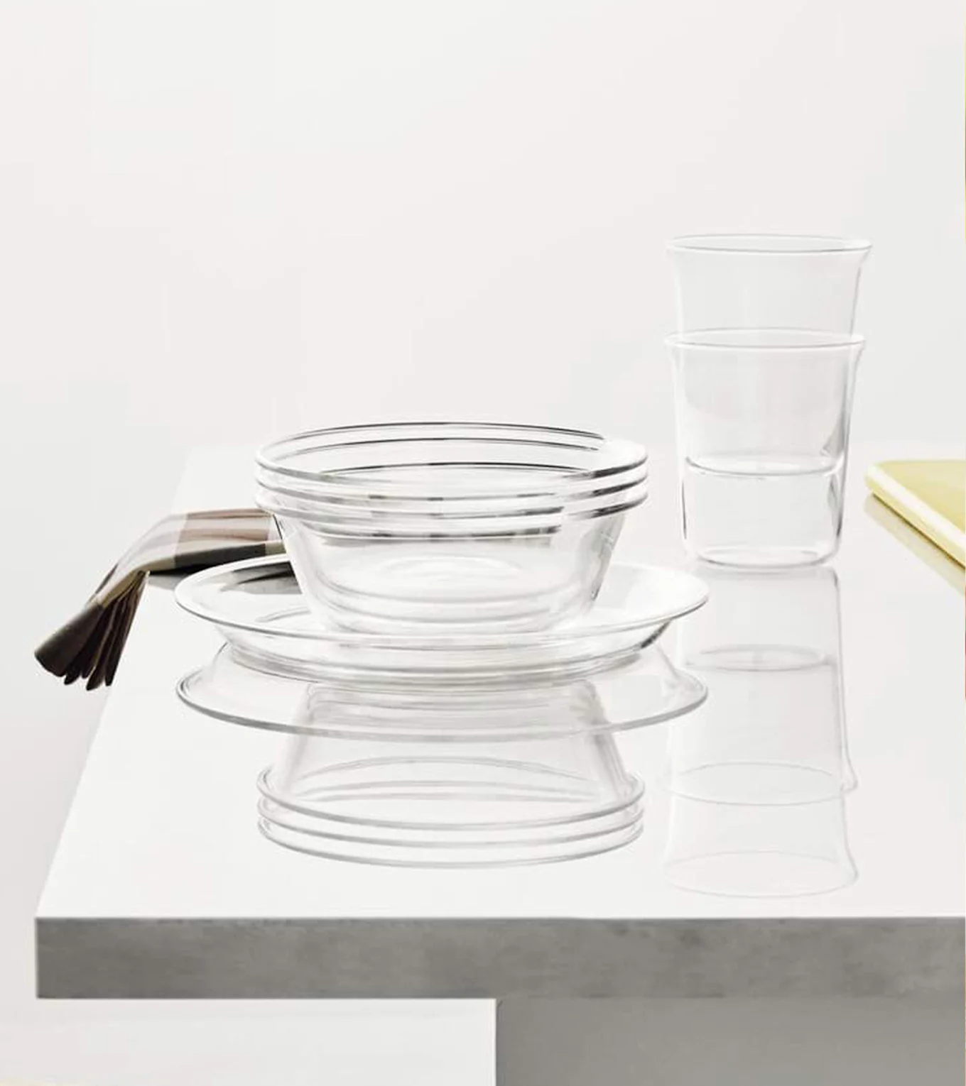 Klare glasskåle, tallerkener og glas stablet på en hvid overflade med en stribet serviet.
