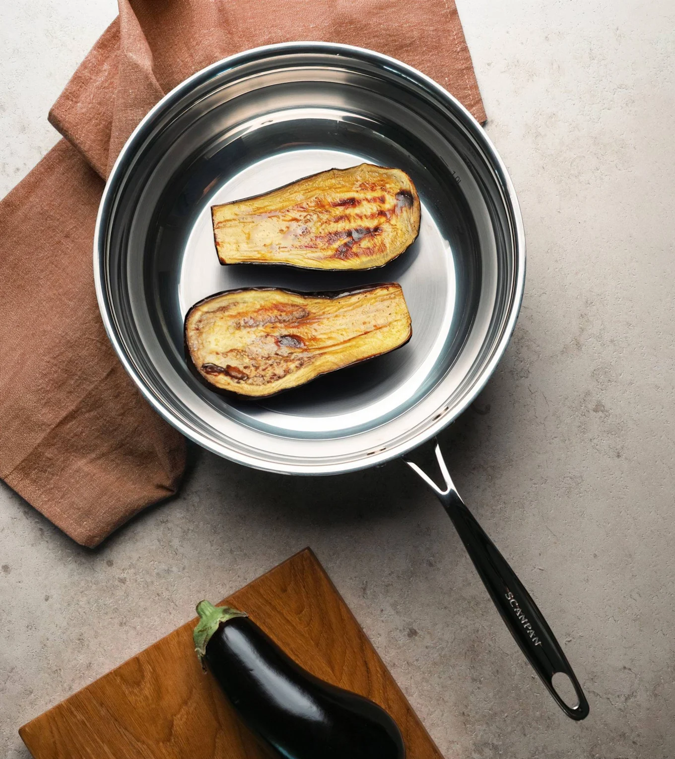 To grillede auberginehalvdele i en stegepande af rustfrit stål, med en rå aubergine på et skærebræt i nærheden.