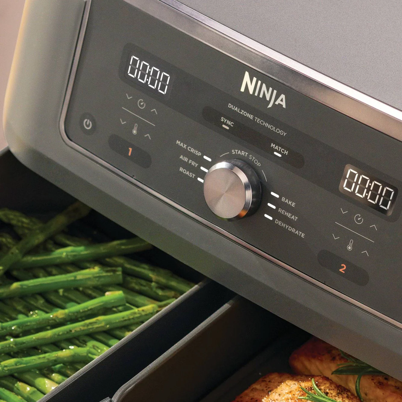 En Ninja airfryers kontrolpanel med to digitale displays og en drejeknap, der viser grønne asparges i den ene skuffe og kylling i den anden.
