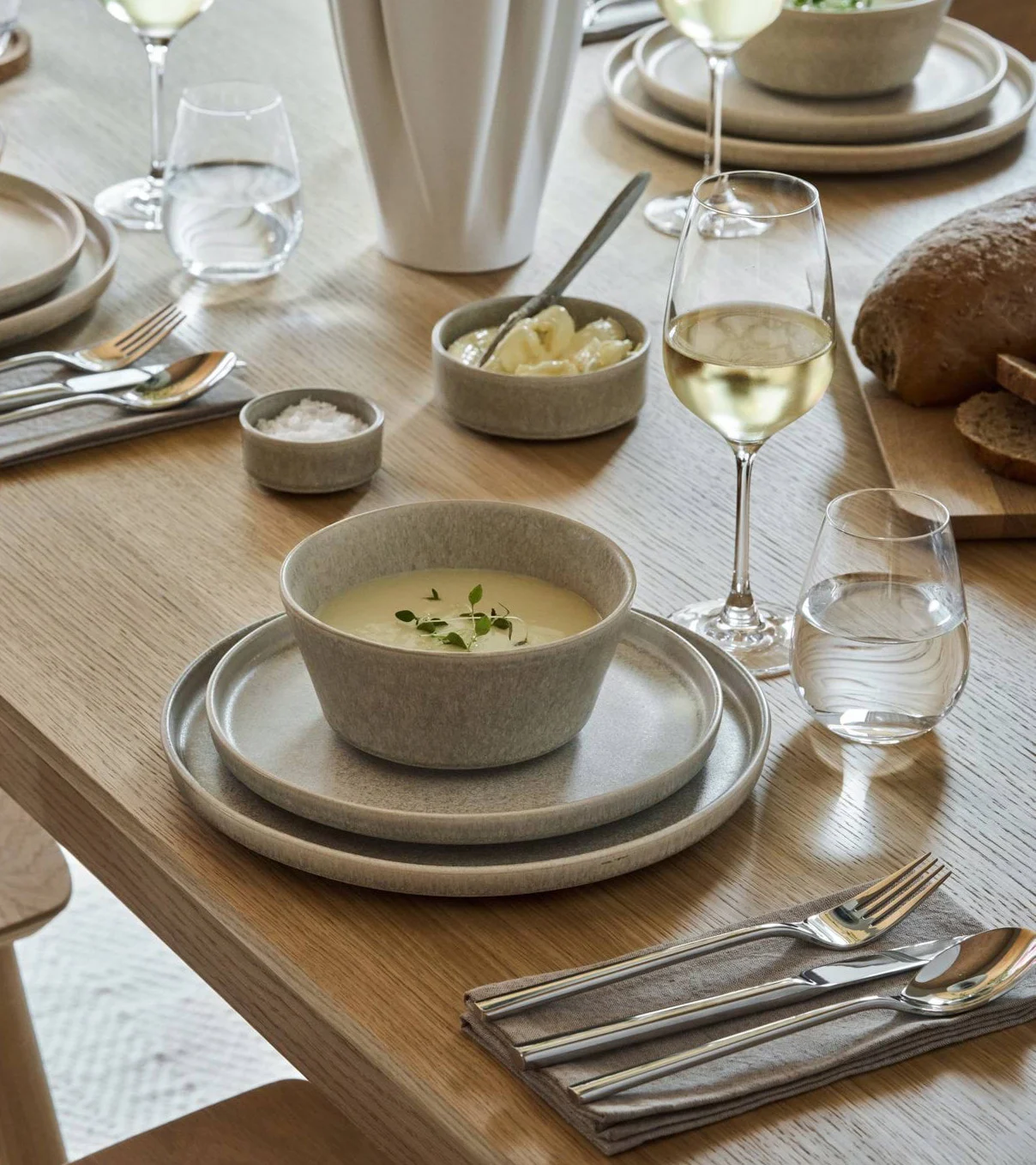 Et spisebord dækket med lysegråt stentøj, suppe, brød, vin, vand og bestik.