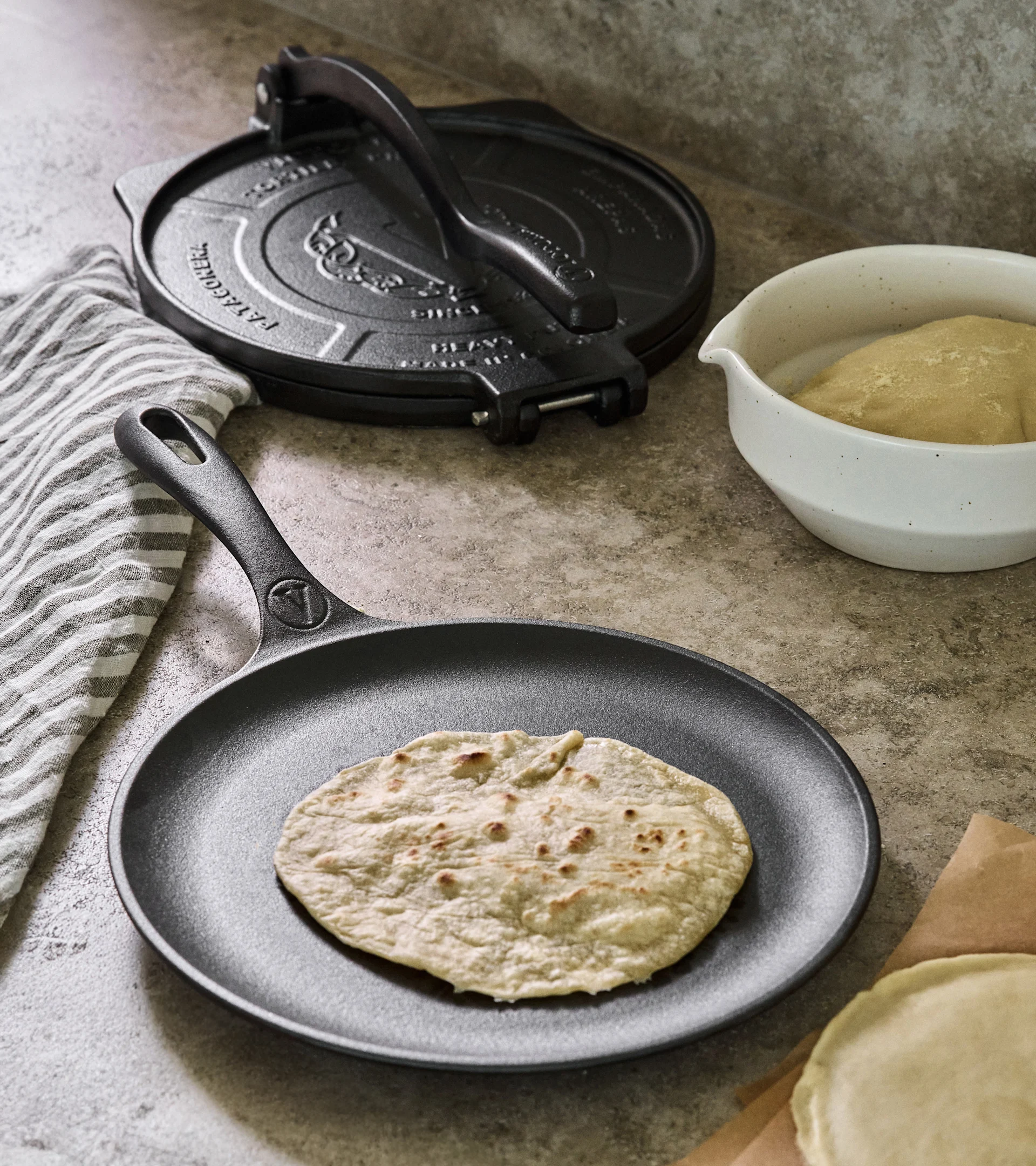 Tortillapresser og stegepande i støbejern med stegt tortilla, dej og rå tortillas på en bordplade.