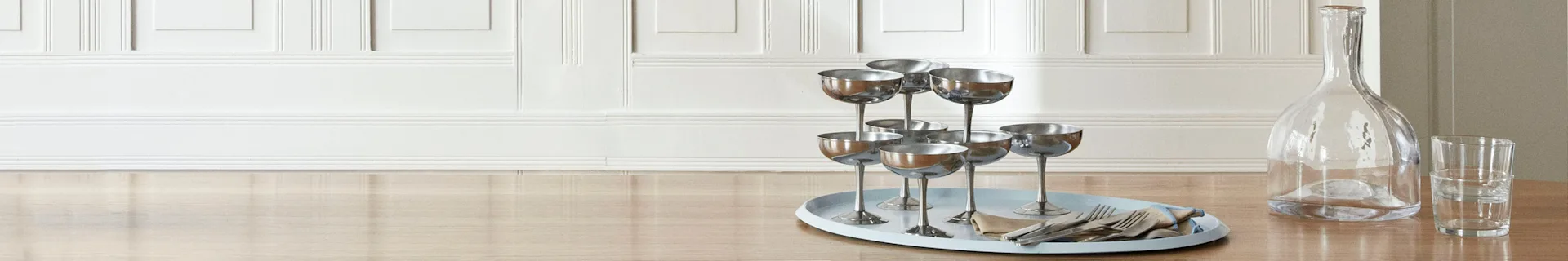 En lyseblå oval bakke holder sølv champagneglas, bestik og en serviet på et lyst træbord ved siden af en glaskaraffel.