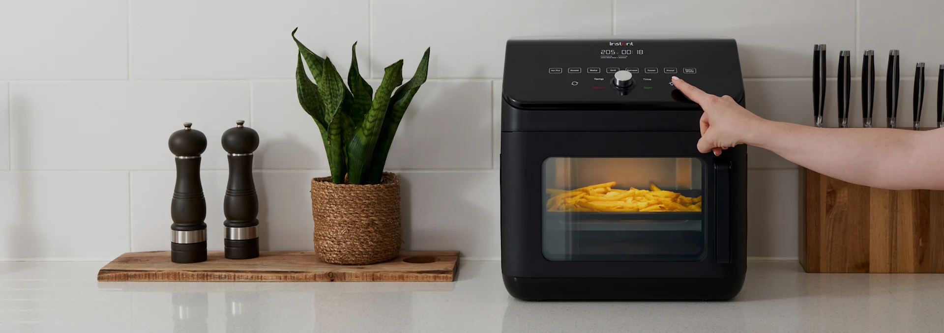 Hånd trykker på knap på sort airfryer med pommes frites, ved siden af krydderikværne, en plante og knivblok på køkkenbord.