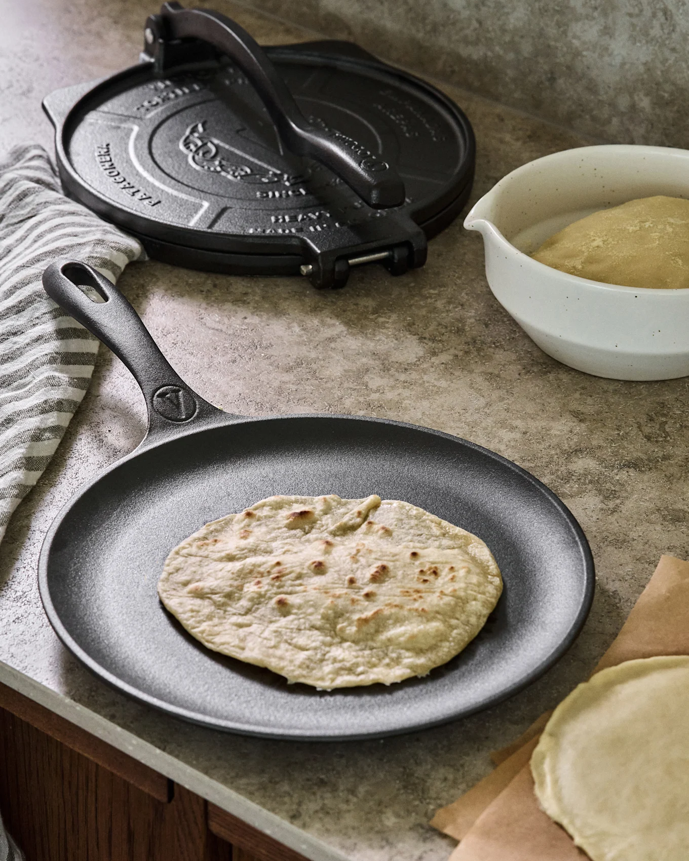 En tilberedt tortilla hviler på en sort støbejernsstegepande, med en tortillapresse, dejskål og ubagt tortilla i baggrunden.