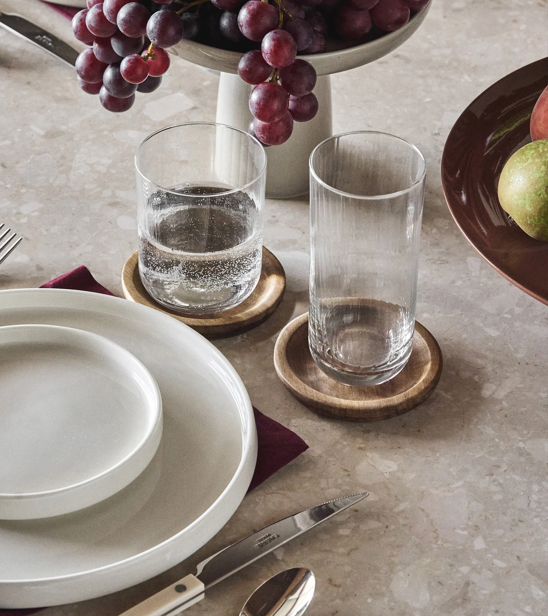 Spisebord med hvide tallerkener, to klare glas på træbrikker, røde druer og et stykke frugt.