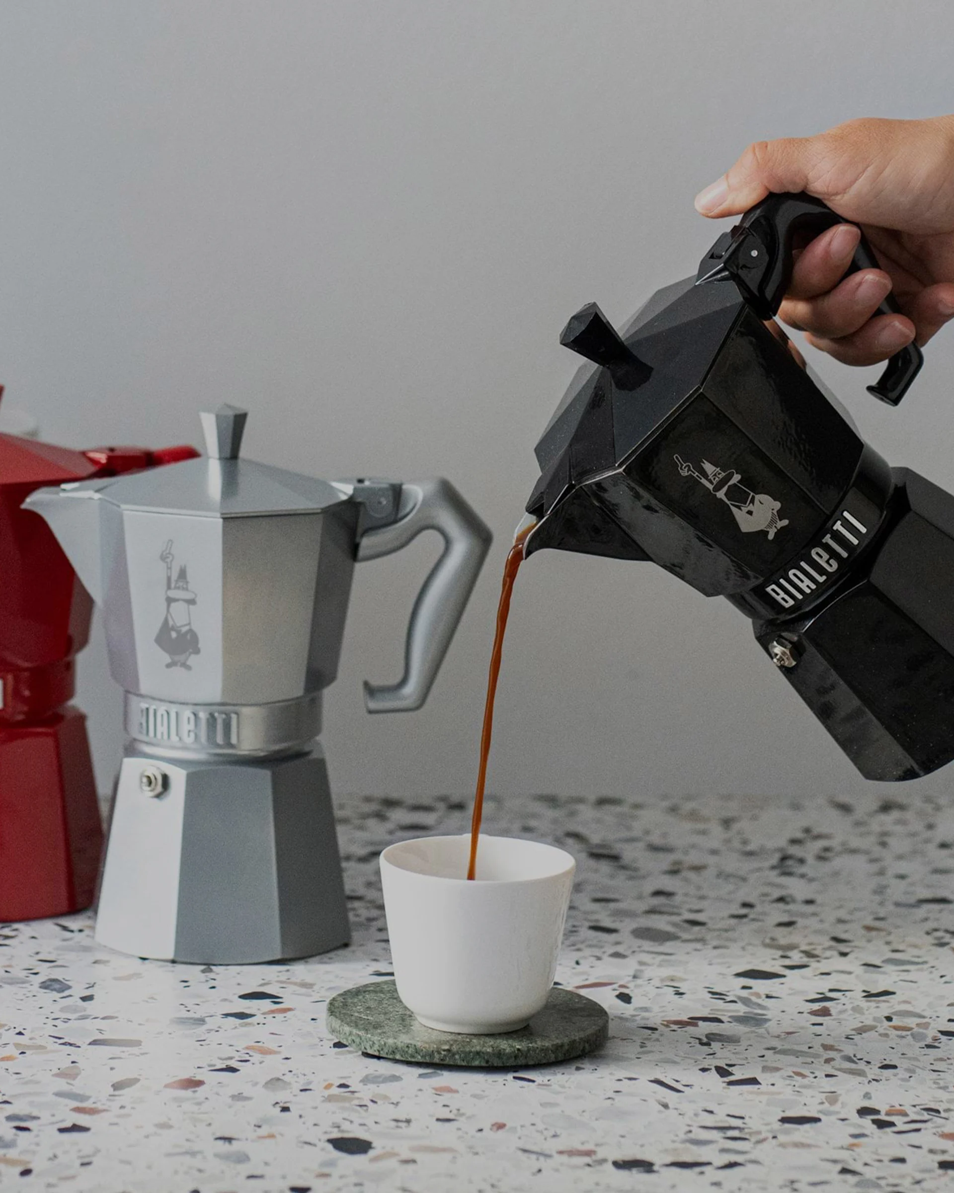 Hånd hælder kaffe fra sort mokkakande i hvid kop, med sølv og rød mokkakande i baggrunden på spraglet bordplade.