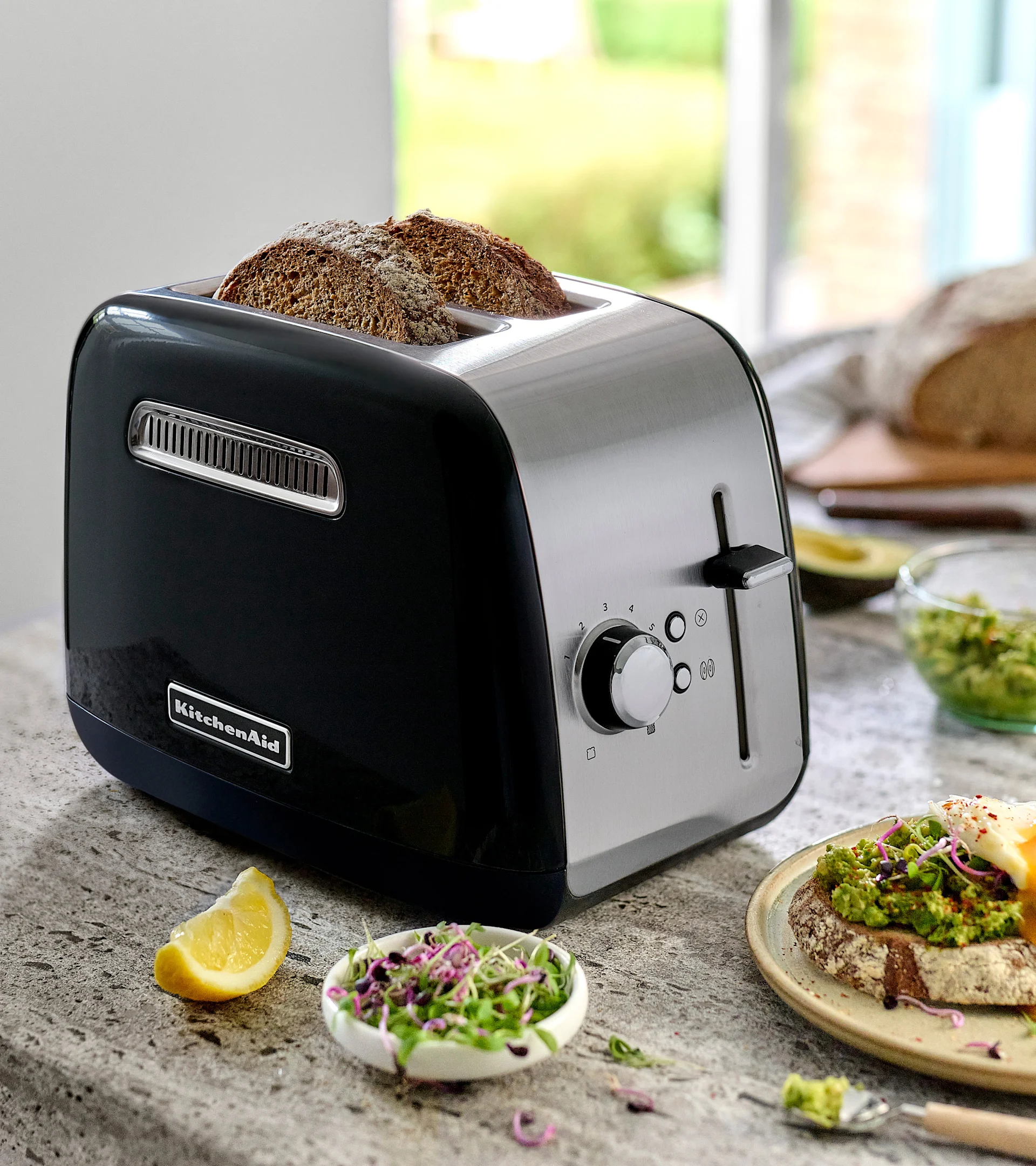 En sort og rustfri stål KitchenAid brødrister med to skiver mørkt brød. På disken, avocadotoast med et æg, spirer og en citronskive.