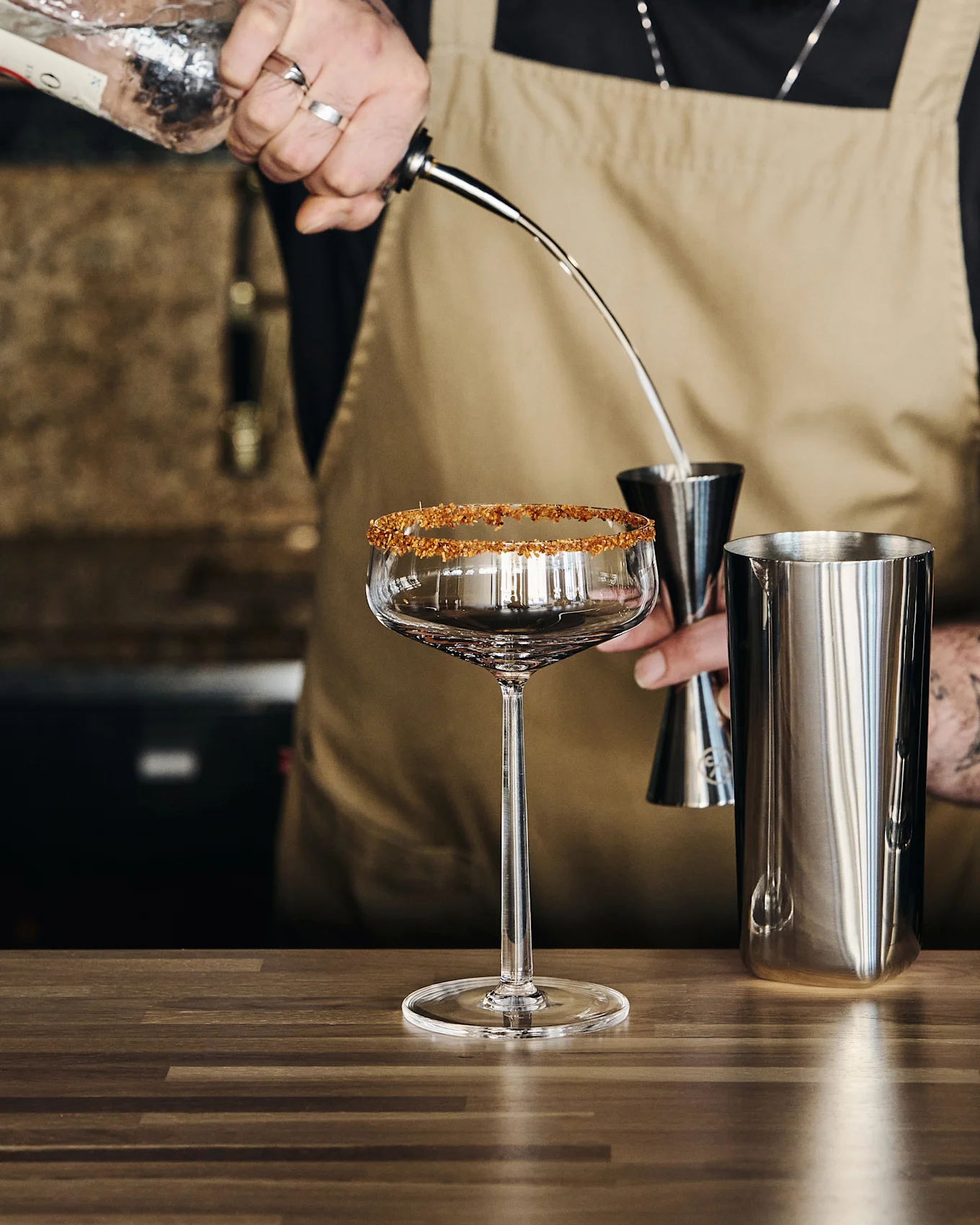 Bartender hælder væske fra en flaske ned i en jigger over en shaker, ved siden af et stilkglas med sukkerkant.