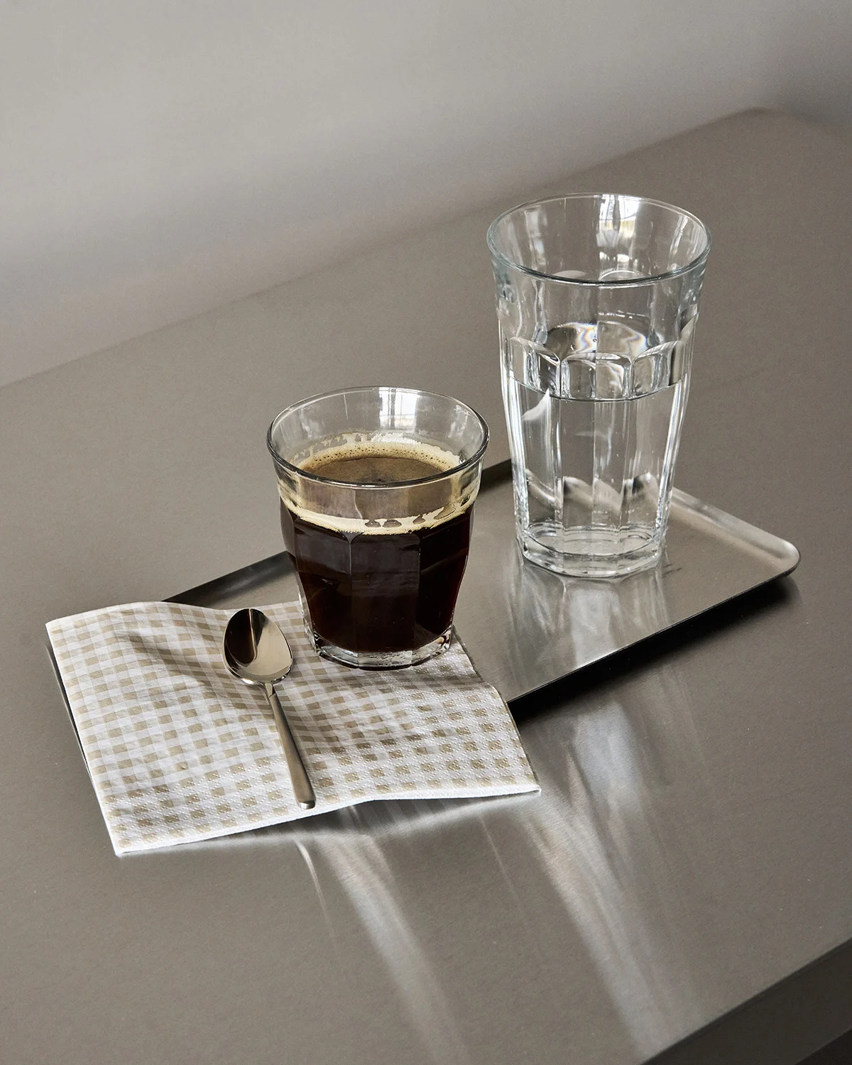 Glas sort kaffe, glas vand og en ske på en ternet serviet på en metalbakke.