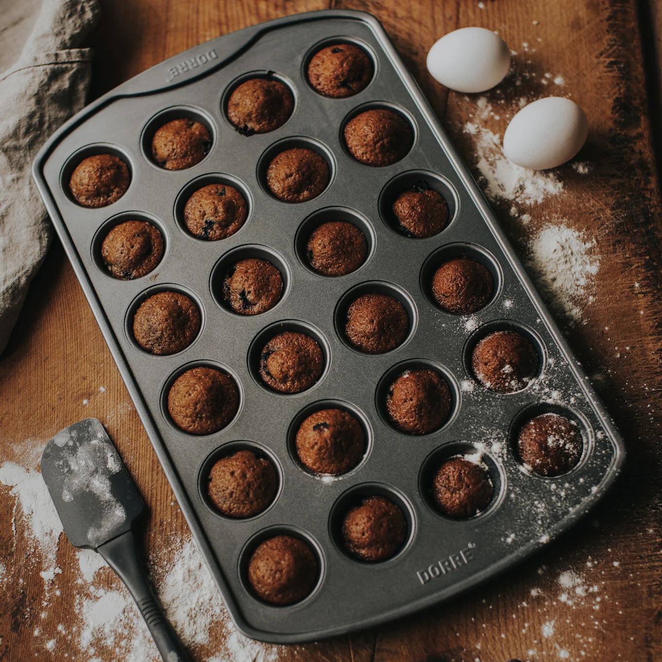 En muffinform med 24 bagte minimuffins på en træoverflade med mel, to hvide æg og en spatel.