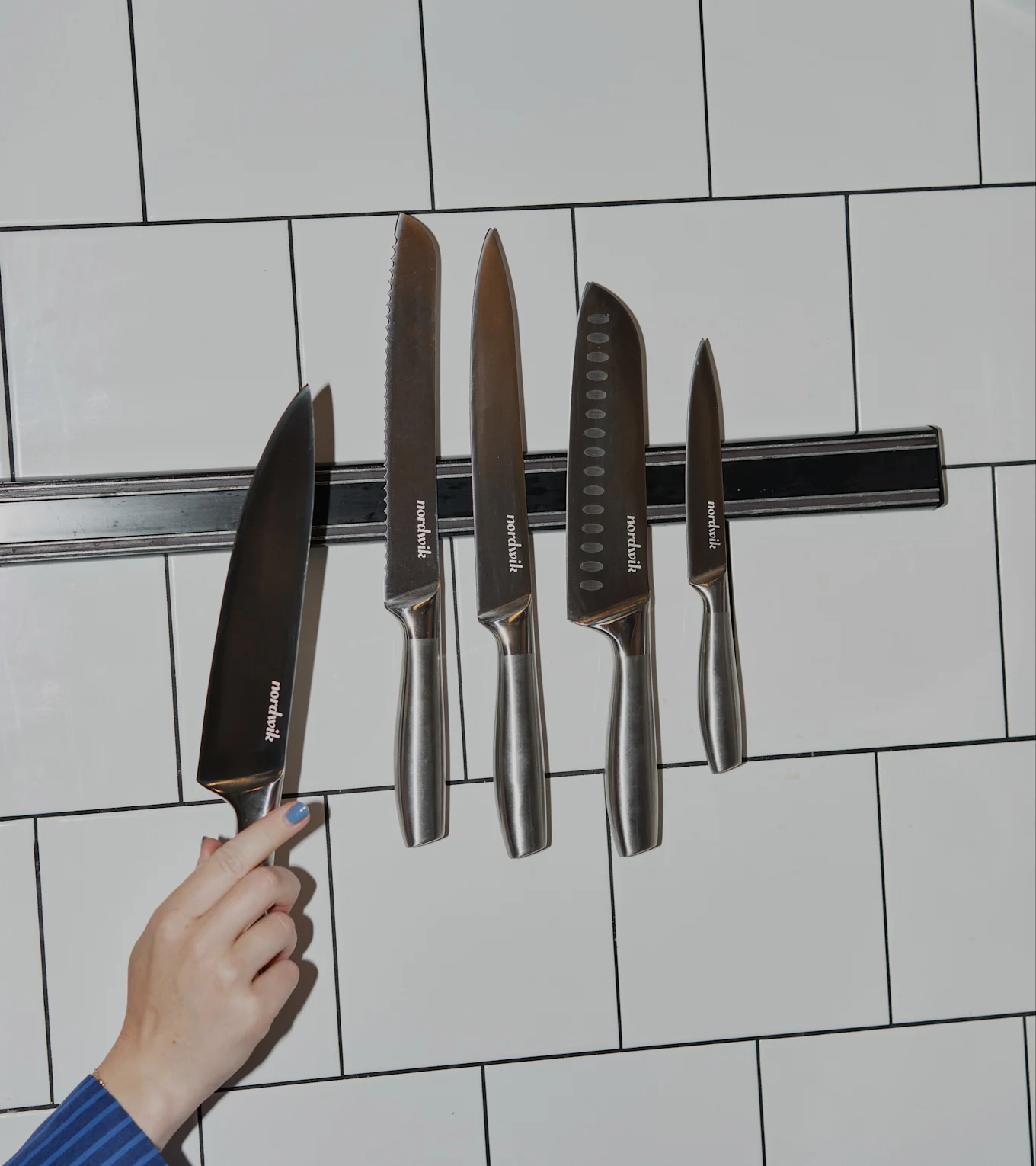 En hånd med blå neglelak holder en Nordwik kokkekniv ved en magnetliste med fire andre Nordwik knive på hvid flisevæg.