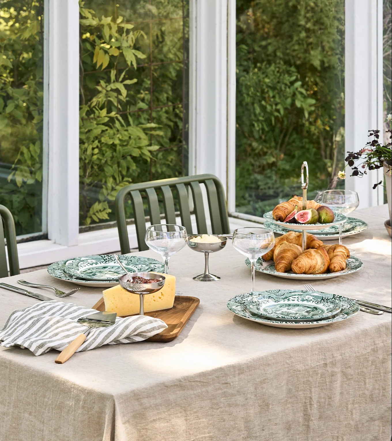 Et morgenmadsbord med grønne mønstrede tallerkener, croissanter, figner, ost, syltetøj og smør i en lys udestue.