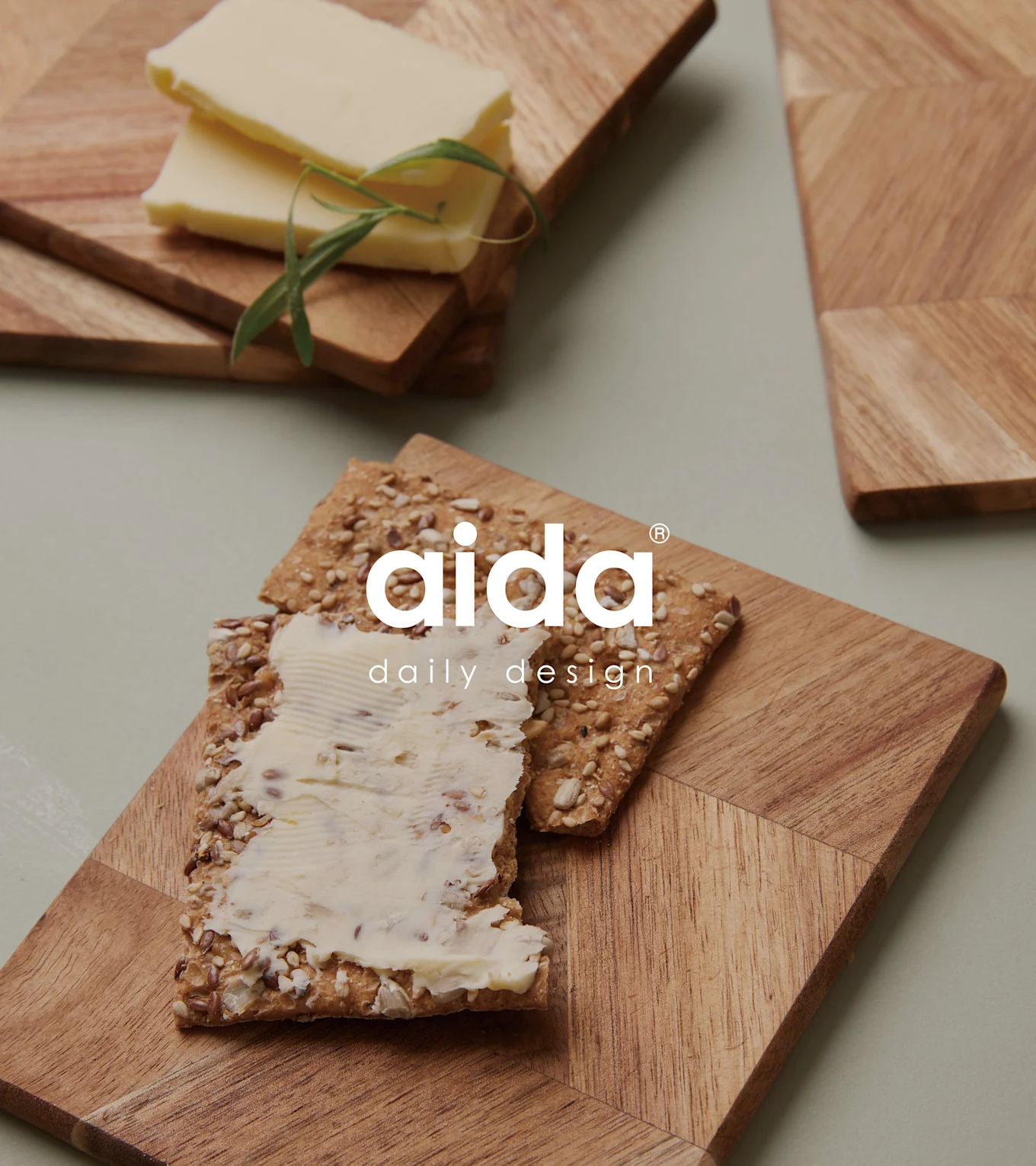 Træbrætter med geometriske mønstre. Det ene holder en kiks med smør, det andet smørskiver og en urt. Aida-logo.