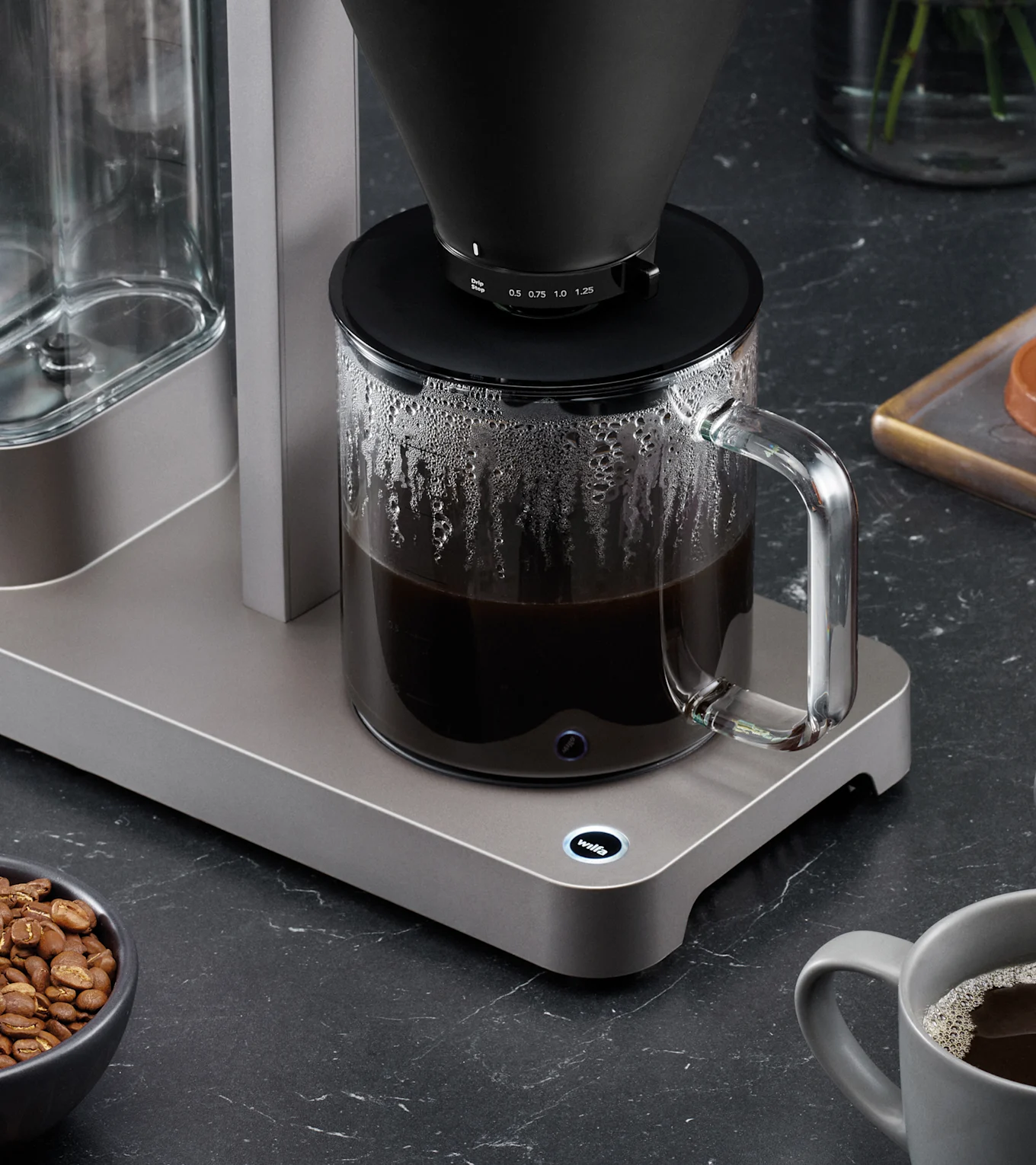 Moderne kaffemaskine med sort filterkegle brygger kaffe i glaskande på mørk bordplade, med kaffebønner og krus.