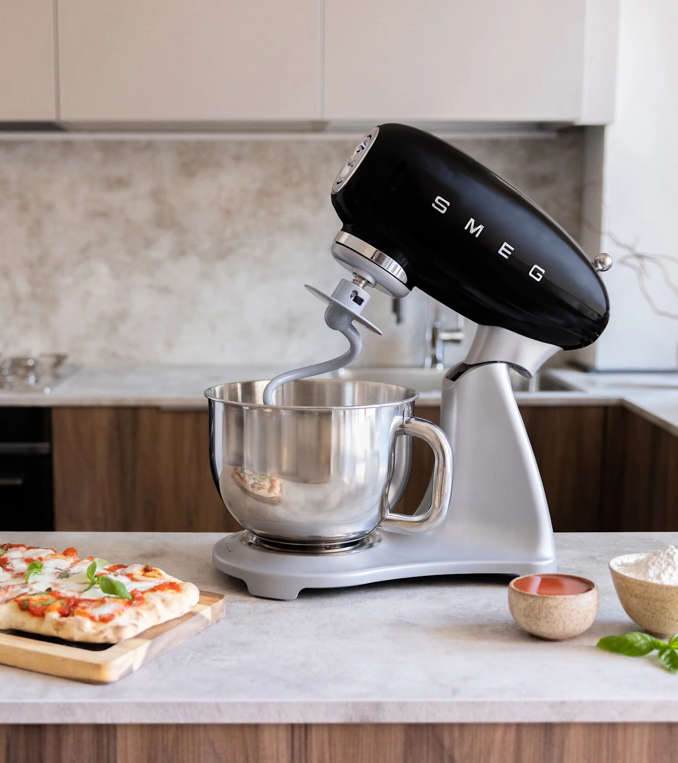 Sort standmixer med røreskål i rustfrit stål og dejkrog på køkkenbord med pizza, mel og tomatsauce.