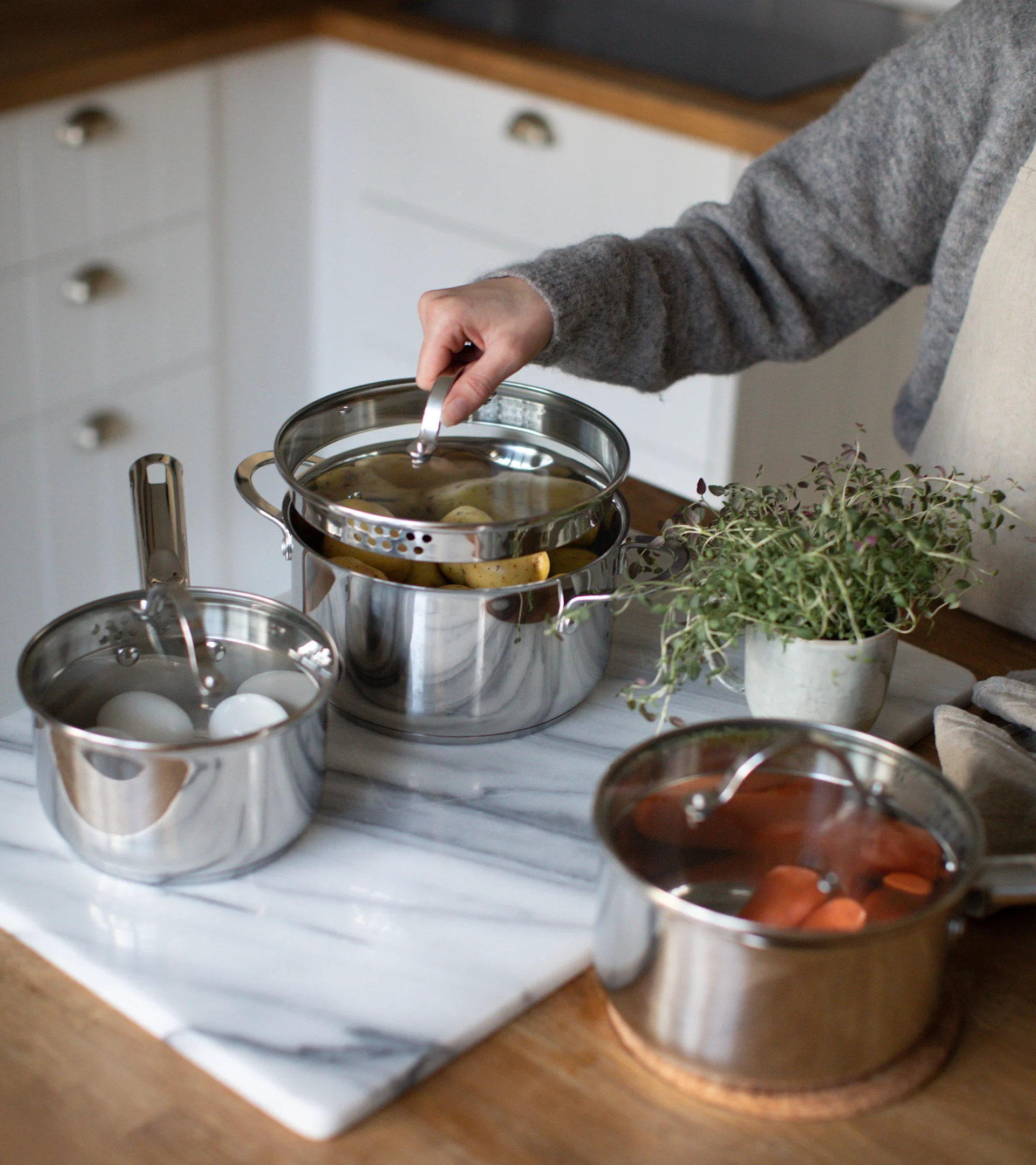 Hånd løfter låg fra en dampkoger i rustfrit stål med kartofler, æg i en anden gryde og gulerødder i en tredje.