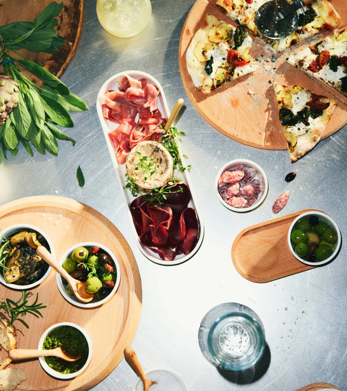 Oversigt over et madbord med en skåret pizza, charcuteribræt, oliven, pesto og tapenade på træfade.