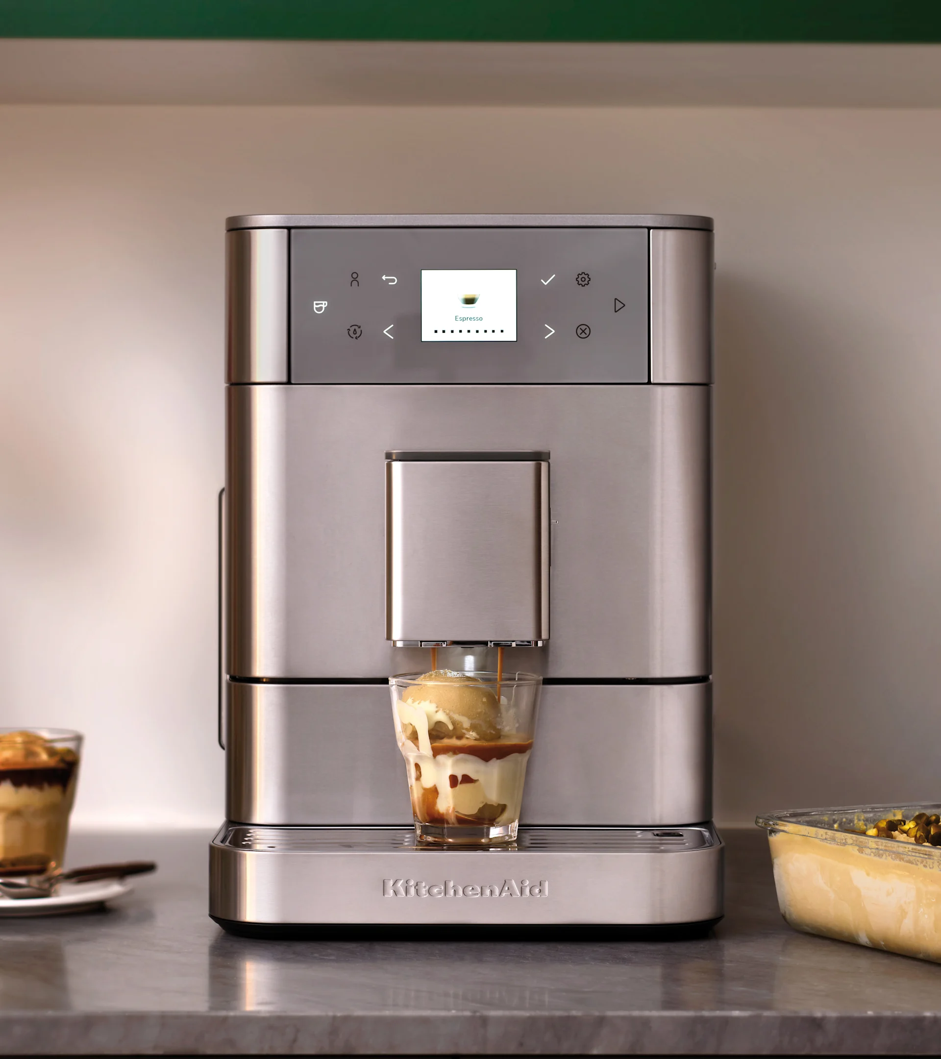 Sølvfarvet KitchenAid espressomaskine laver affogato i et glas med is. Anden dessert i baggrunden.